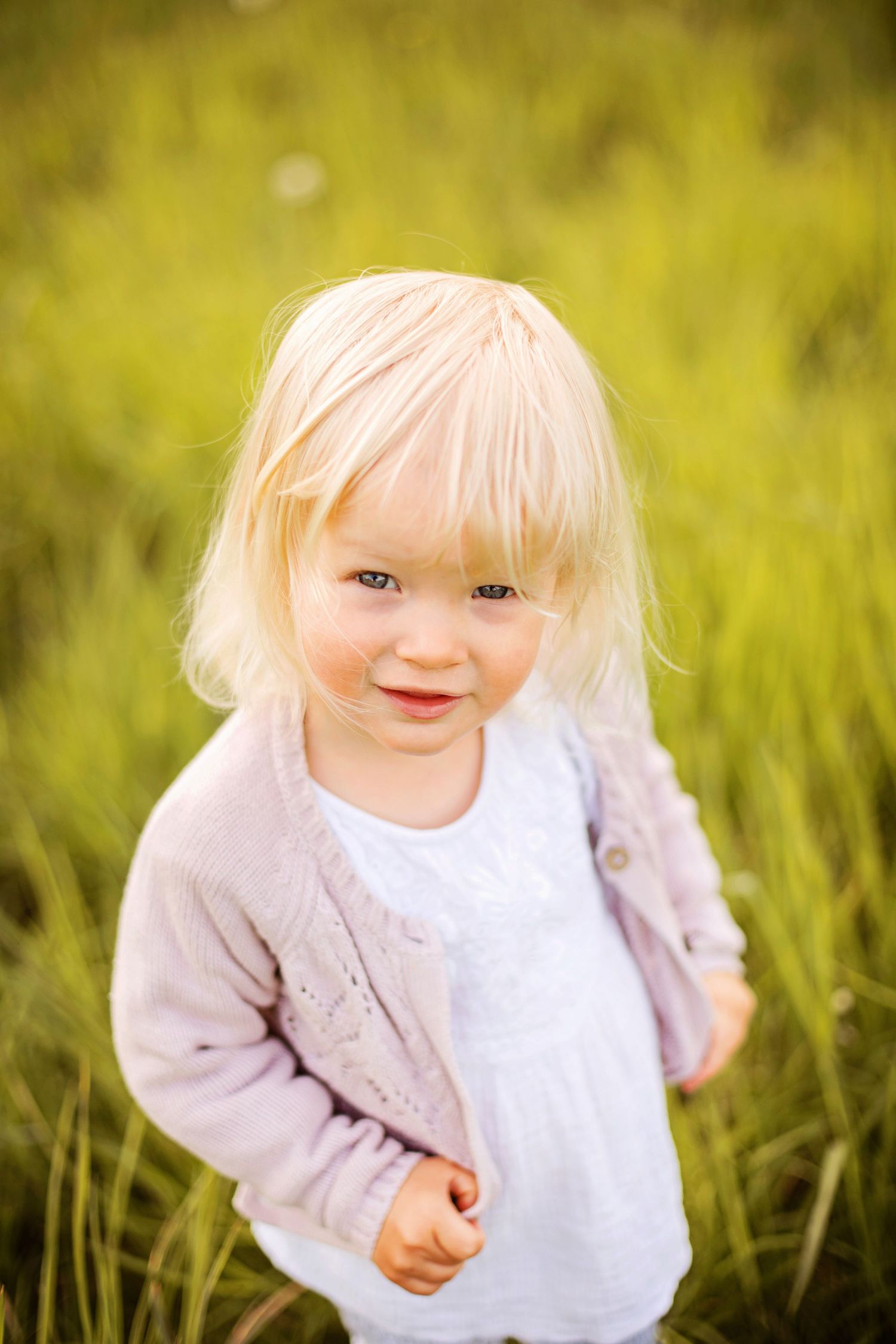 Adorable blonde toddler girl smiling in a green grass field, wearing a white dress and lavender cardigan.