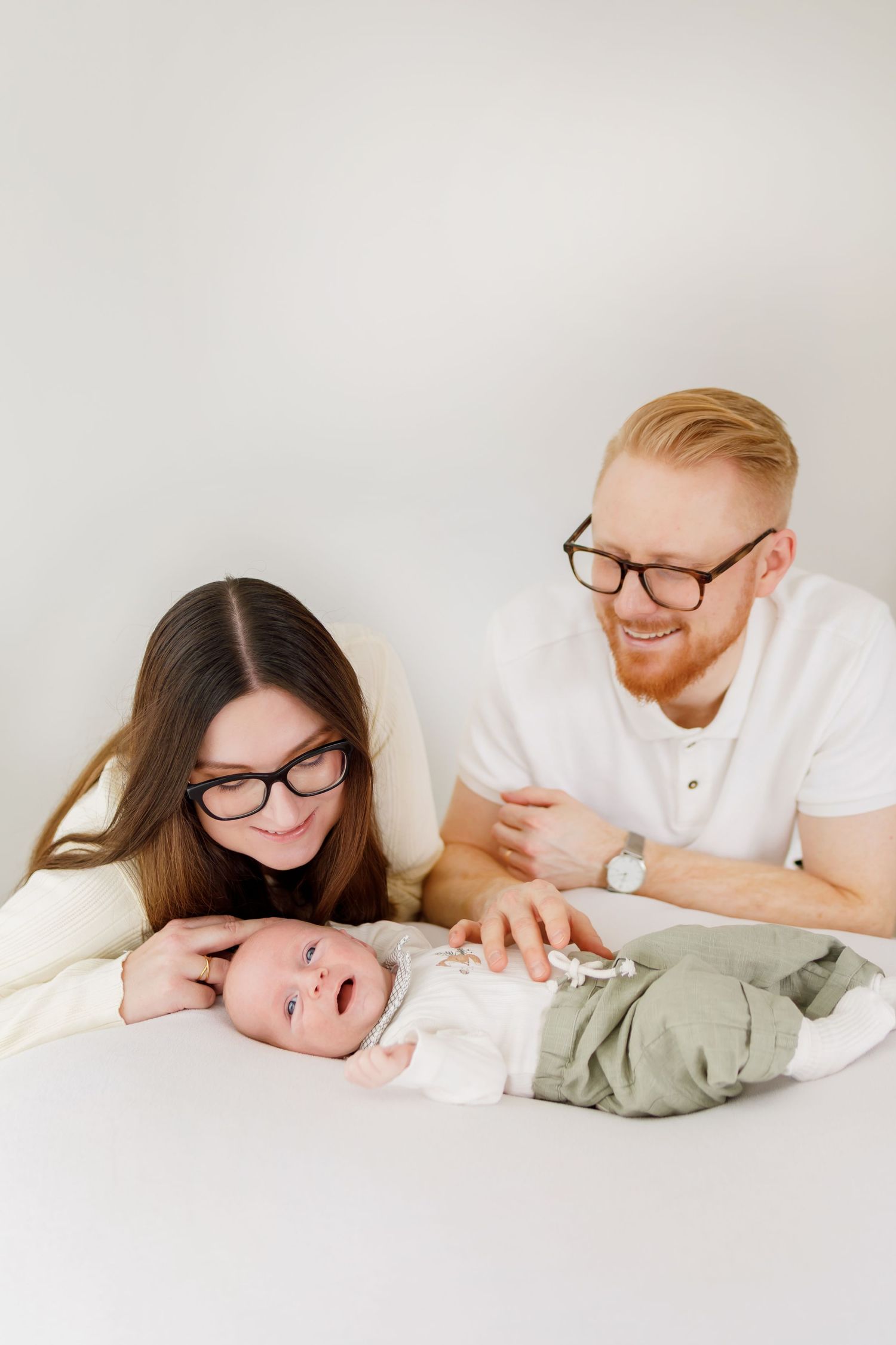 Happy parents with glasses smiling at their newborn baby lying on a white surface.