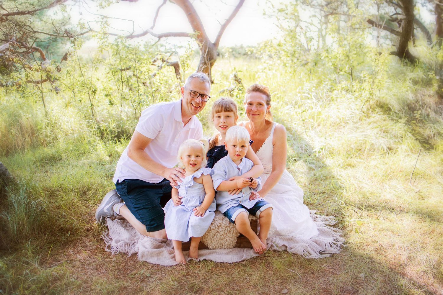 A family group sits together outdoors in a sunlit wooded setting wearing coordinated white outfits.