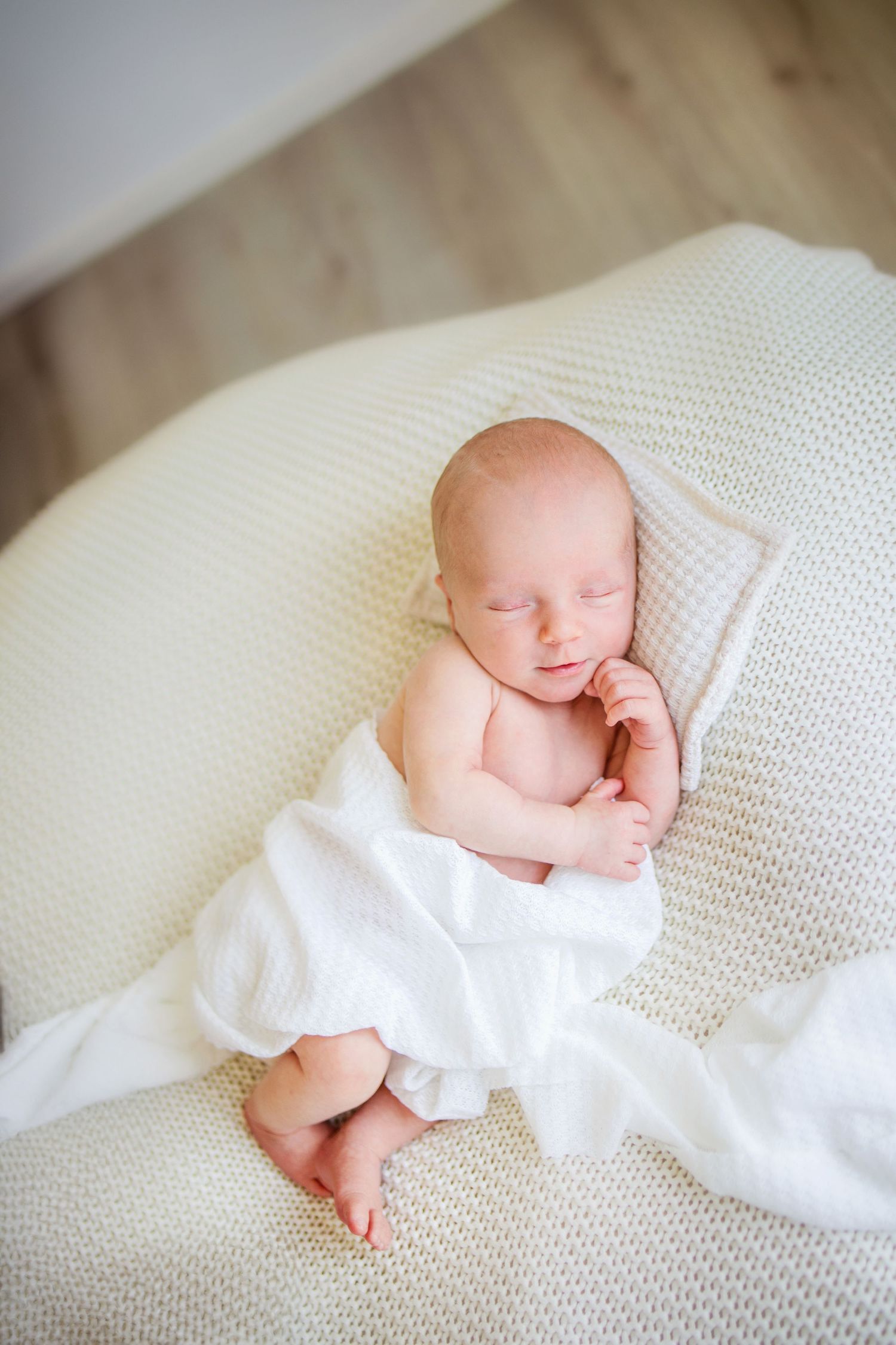 A peaceful sleeping newborn swaddled in soft white fabric rests on a textured cream-colored blanket.