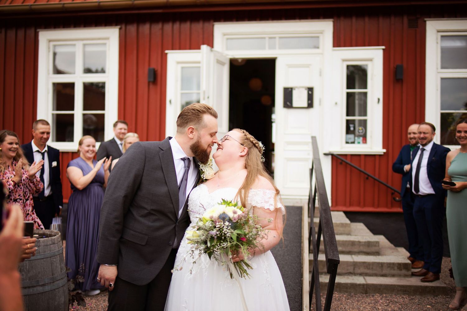 Newlywed couple sharing a kiss outside a red Swedish venue, surrounded by applauding wedding guests.