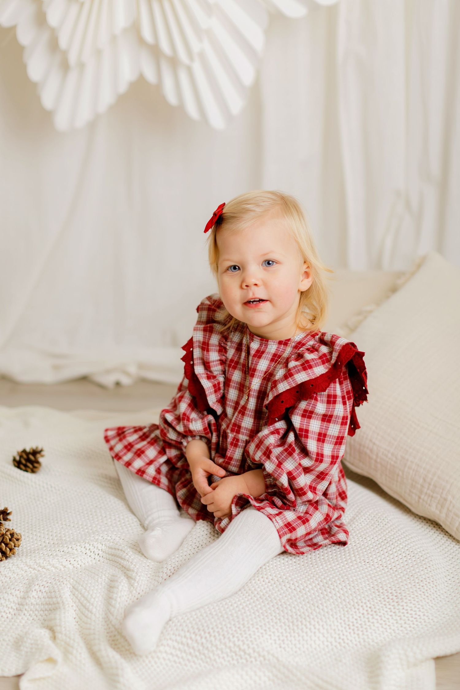 Toddler girl in red plaid dress with bow sits on white blanket in bright holiday-themed photo setting.