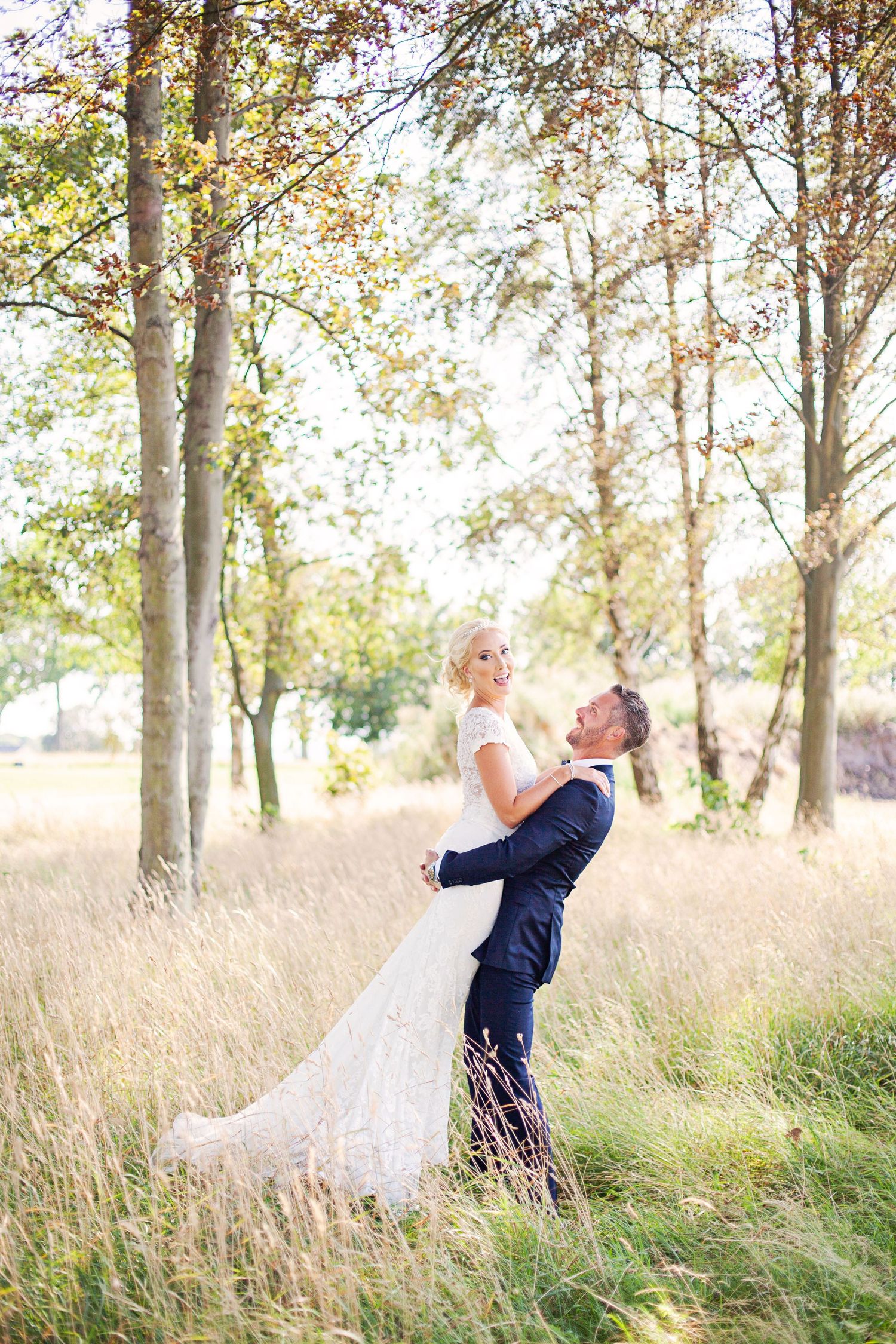 Groom lifting bride in white lace gown outdoors in golden meadow with trees, romantic wedding portrait.
