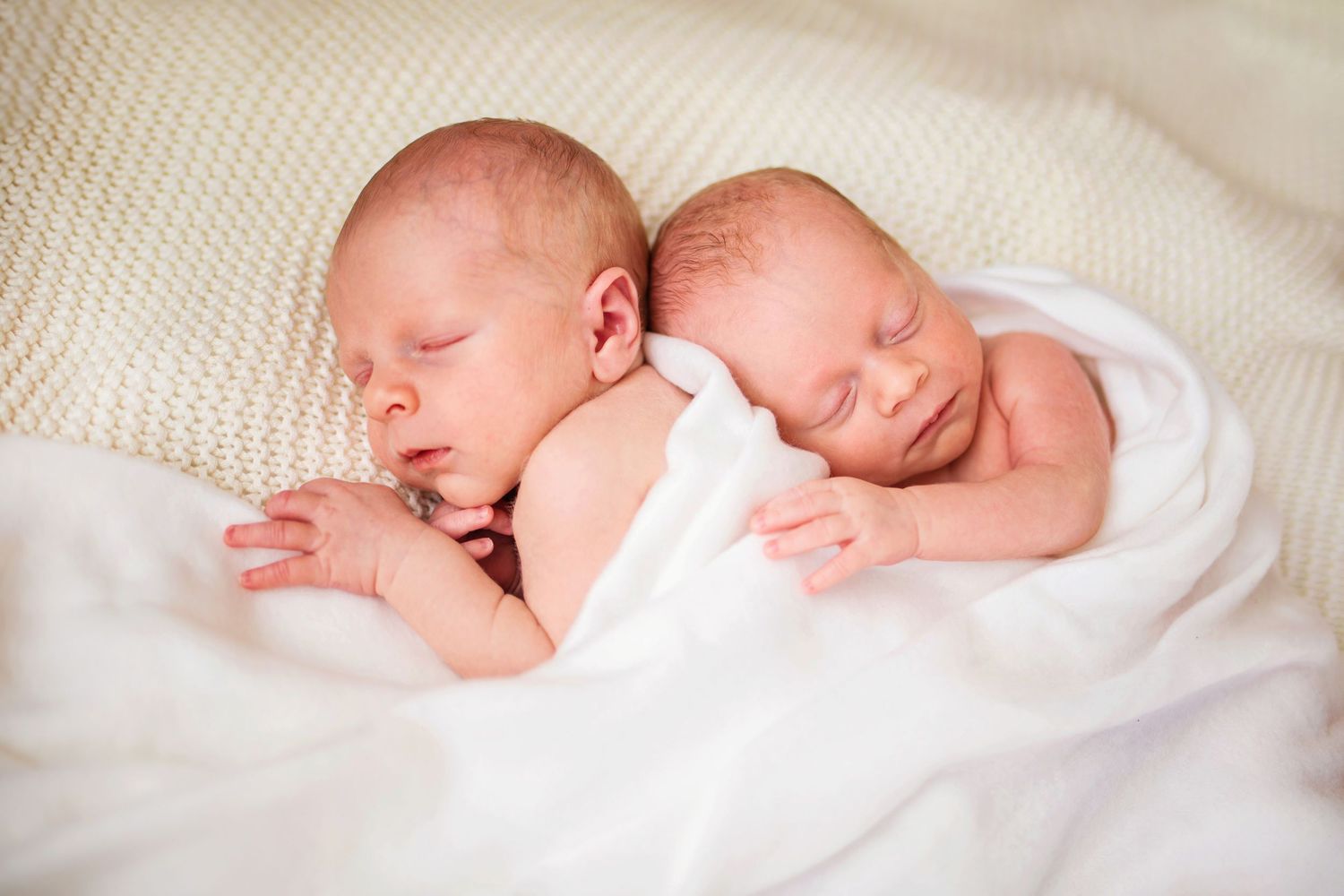 Two newborn twins sleeping peacefully side by side wrapped in soft white blankets.