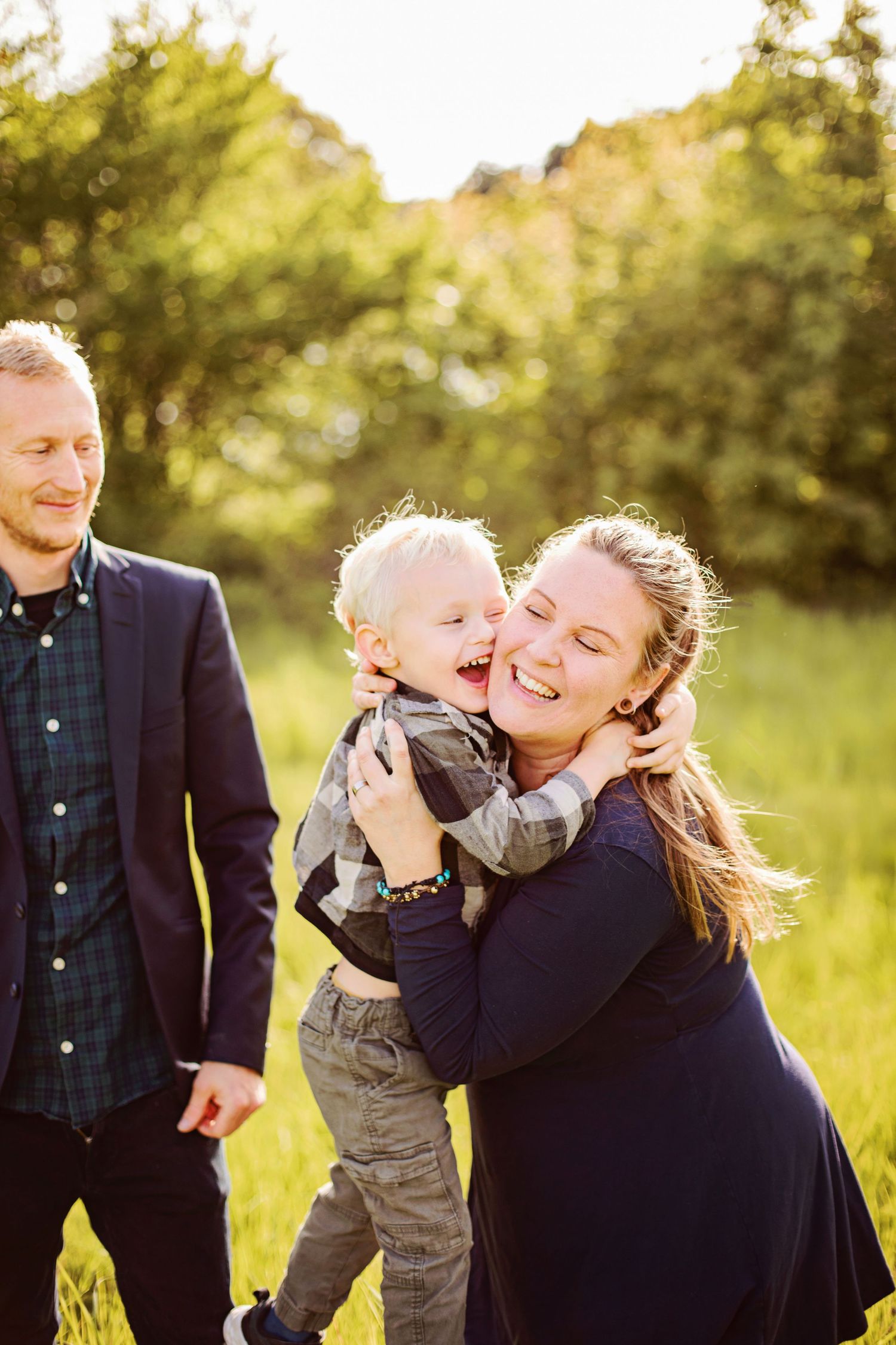A joyful mother hugs her laughing toddler son outdoors while father smiles nearby in a sunny green field.