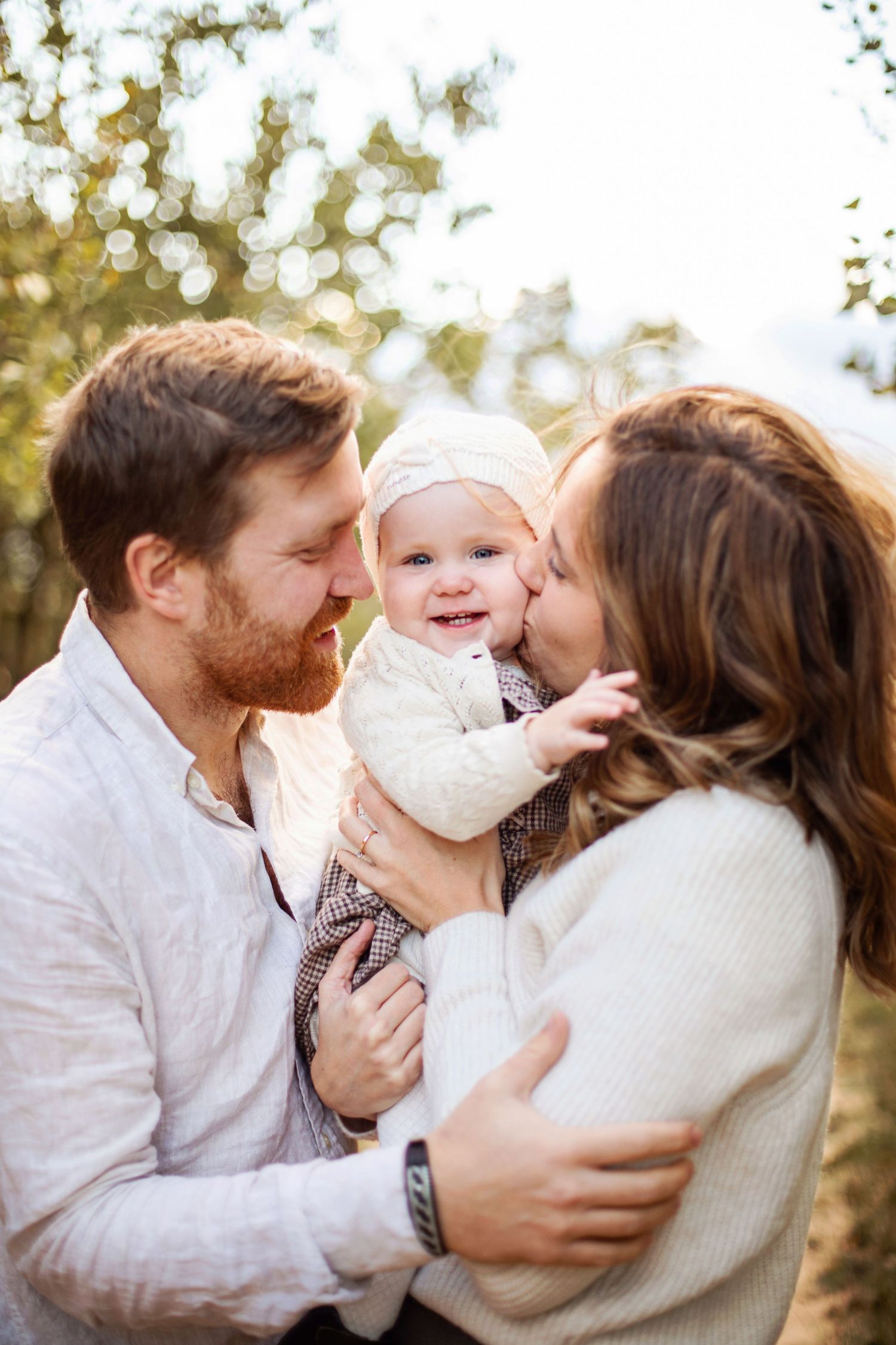 Happy family portrait with parents kissing smiling baby outdoors in soft natural light.