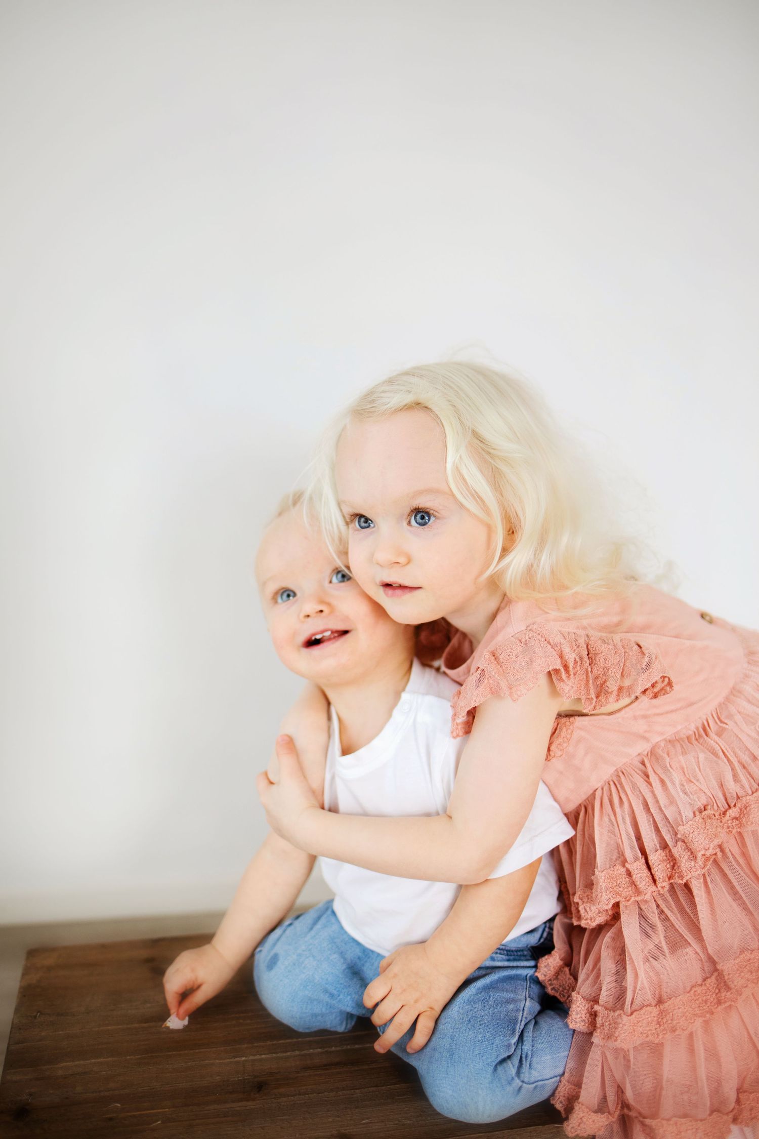 Two blonde toddlers with blue eyes, a girl in pink dress hugging a baby in white shirt and jeans, sitting on wood floor.