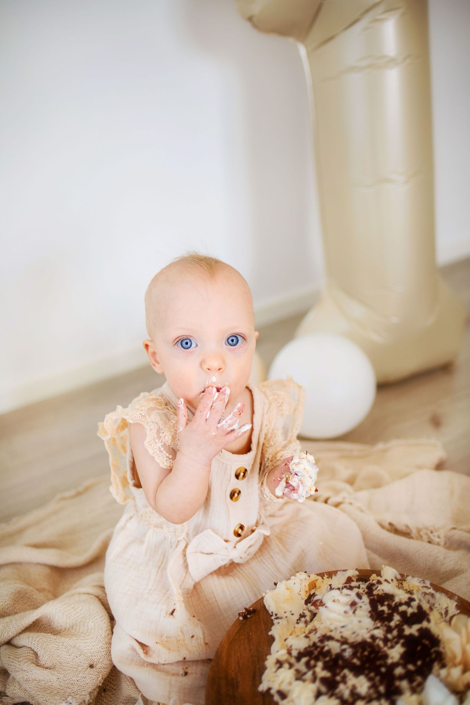 Baby wearing white dress sits on floor with chocolate cake smeared on face during first birthday cake smash photoshoot.