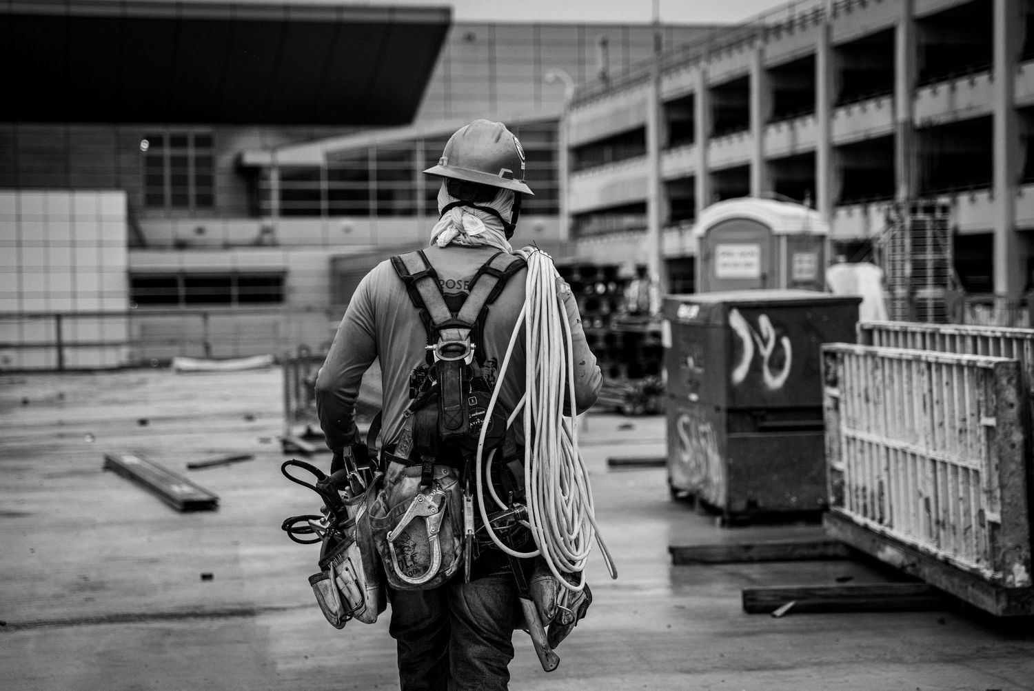 A black and white photo of a construction worker carrying coils of rope and equipment on a building site.