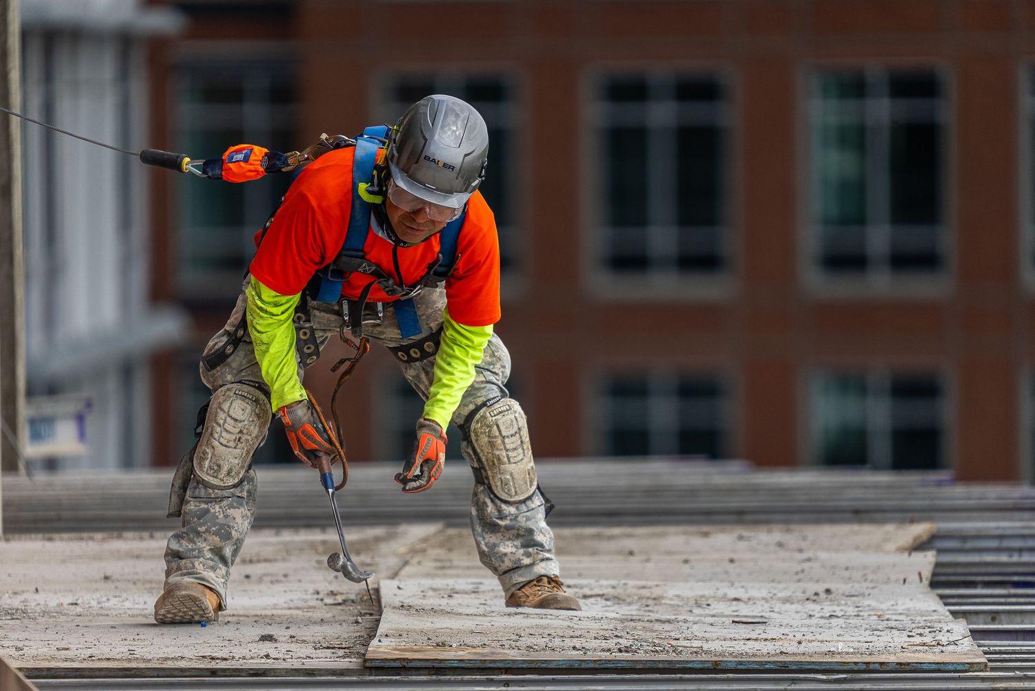 Construction worker in safety gear crouching on a concrete surface with power drill against urban building backdrop.