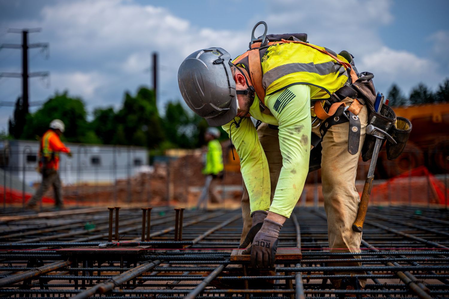 Construction worker in safety gear bends down to work on metal rebar grid at construction site on sunny day.