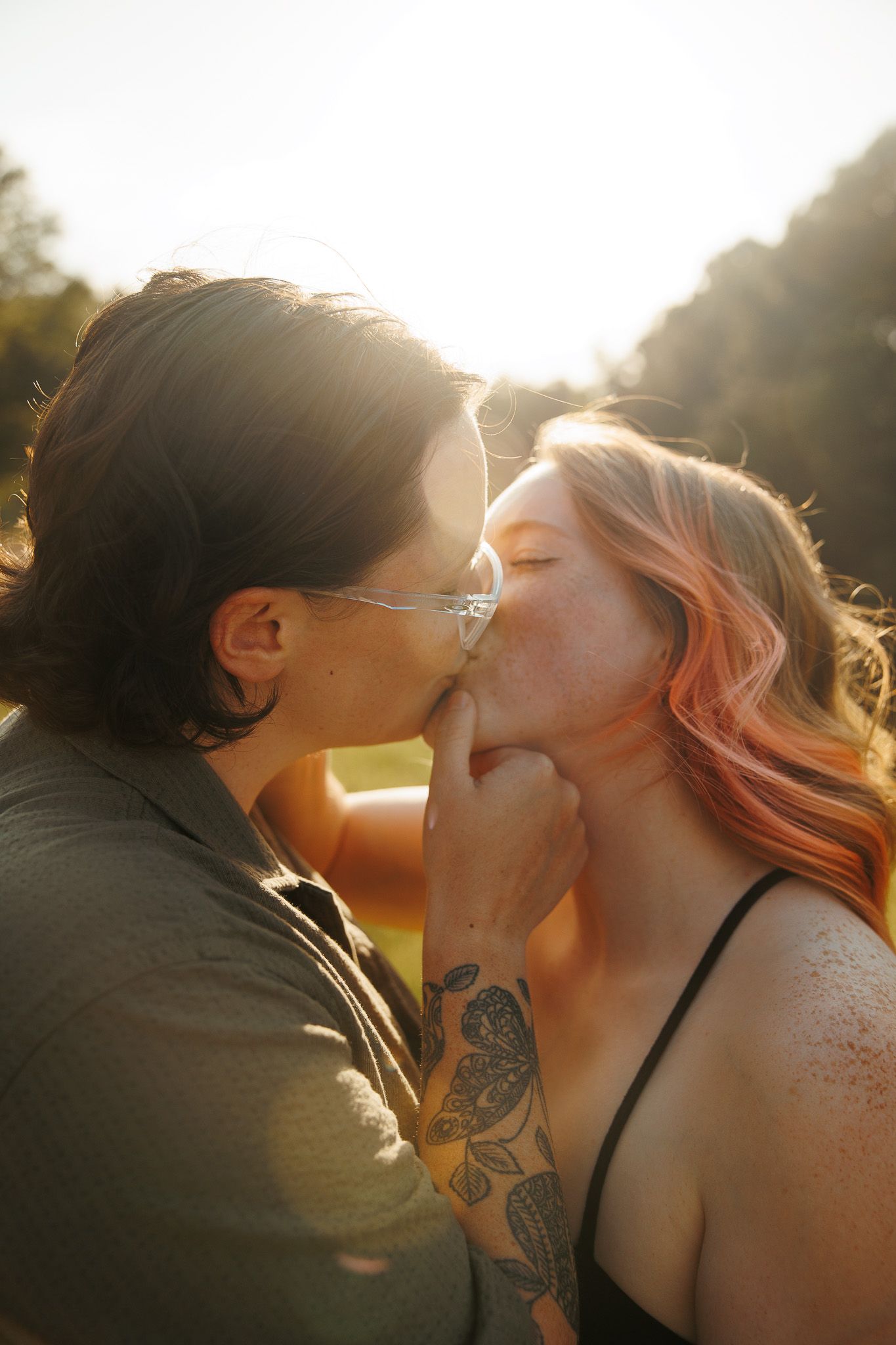 Couple sharing a kiss in warm sunlight, woman with pink hair and floral tattoo, backlit by sun.
