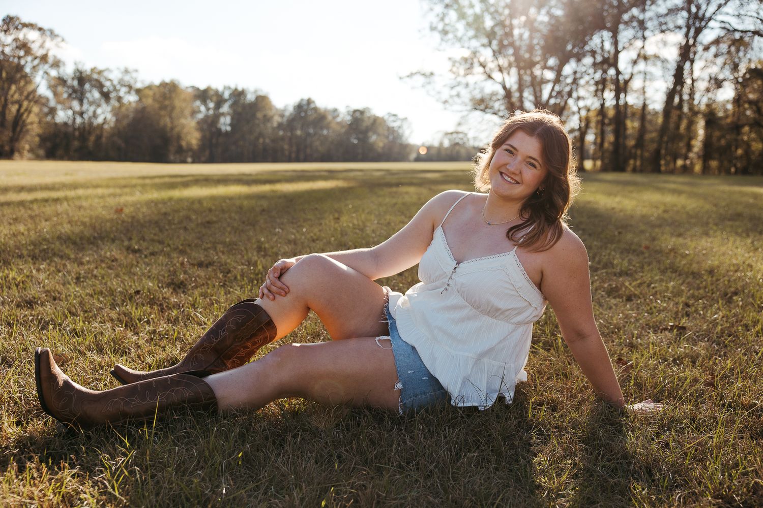 Young woman in white top, denim shorts, and cowboy boots sitting on green grass field with sunlit trees in background.