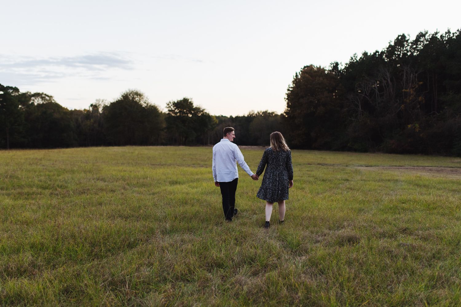 Couple holding hands walking through an open grassy field at dusk with trees in background.