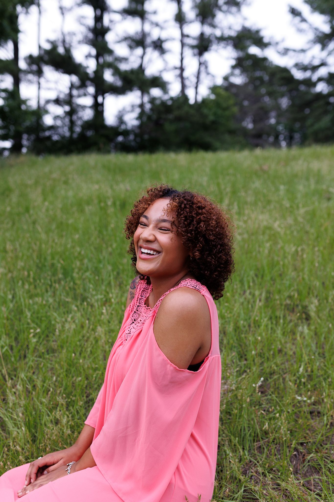 A joyful girl in a pink cold-shoulder dress sits laughing in a lush green field with trees behind her.