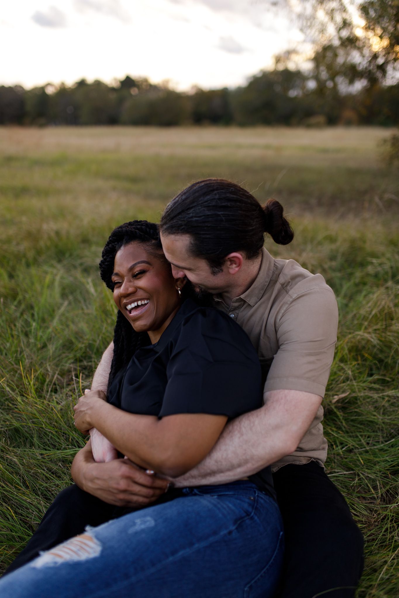 Couple sitting in a grassy field, man embracing woman from behind as she laughs joyfully at sunset.
