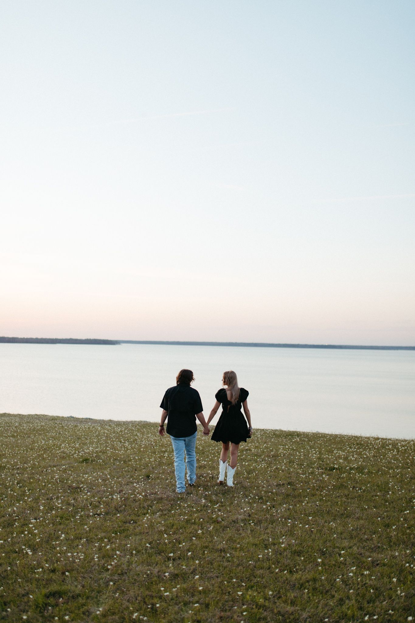 Couple walking hand in hand toward calm lake at sunset, woman in black dress and white boots.