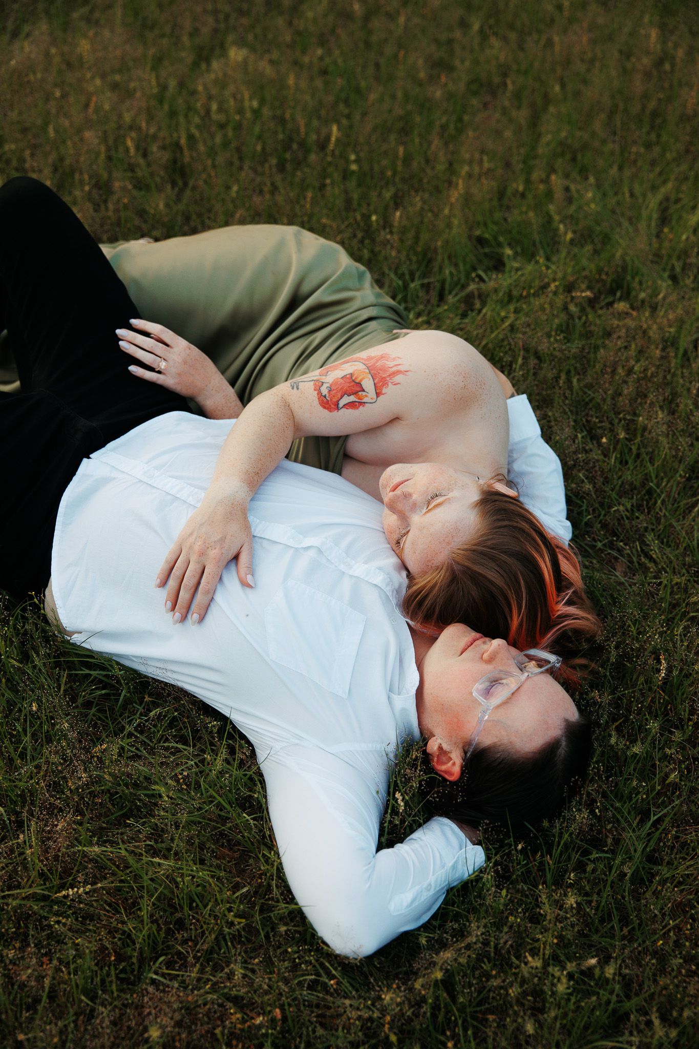 Couple lying together in green grass, woman in sage strapless dress resting on partner's chest.