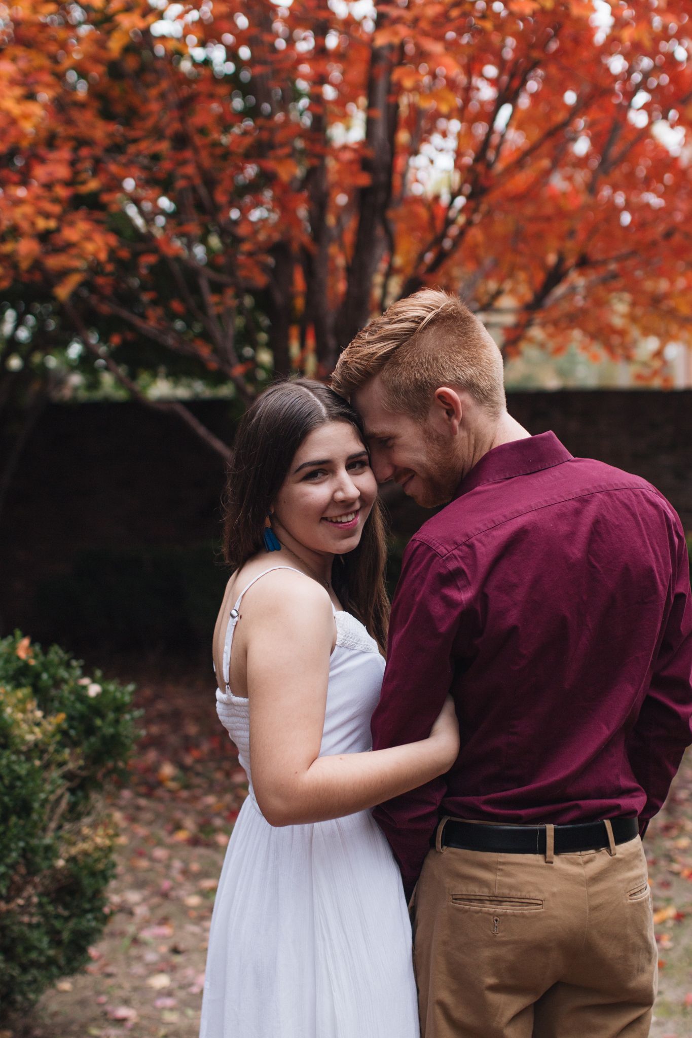 Couple embracing tenderly in front of vibrant red autumn trees, woman smiling at camera.