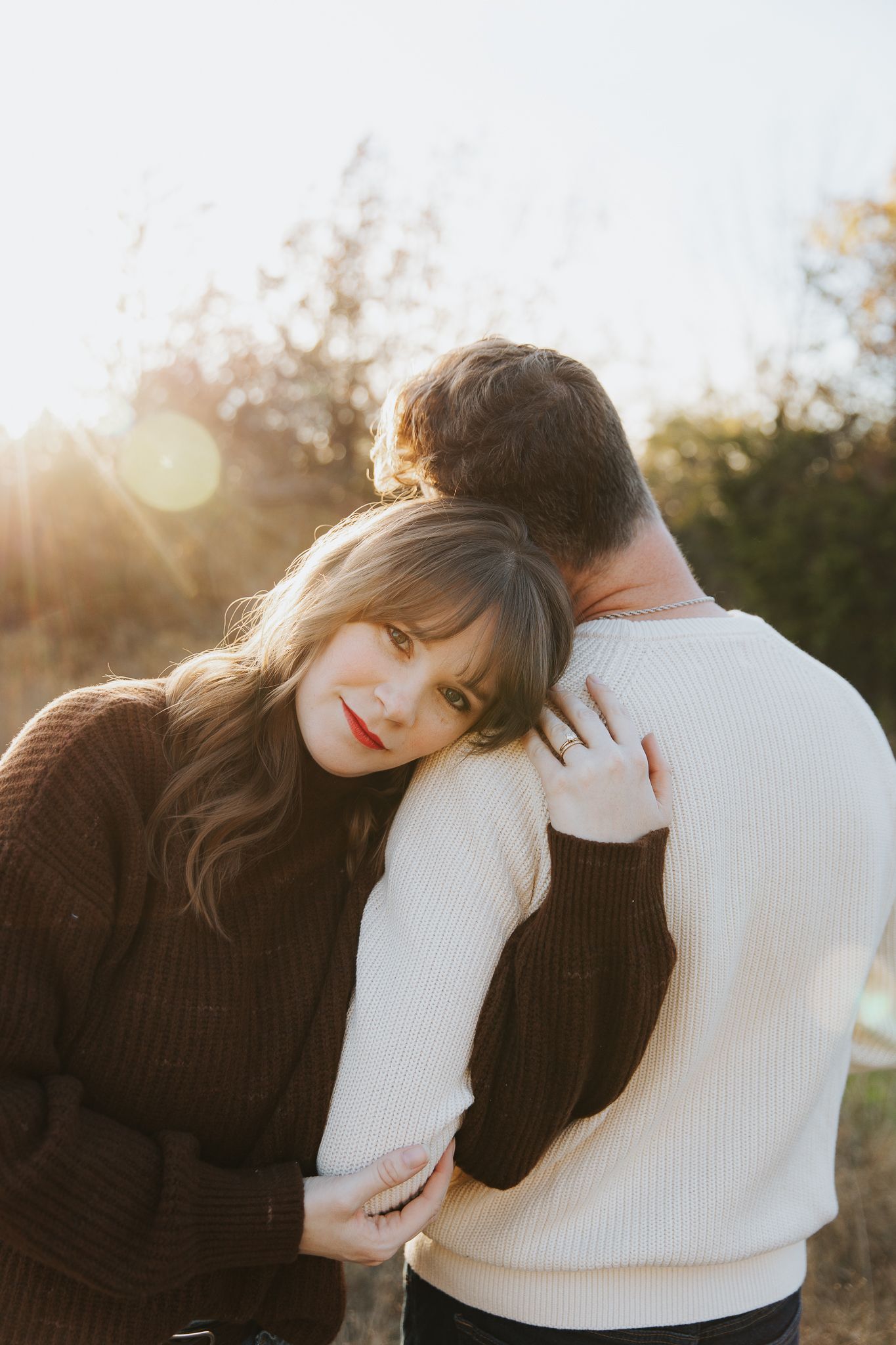 Woman in brown sweater leaning on mans shoulder in warm golden sunset light.