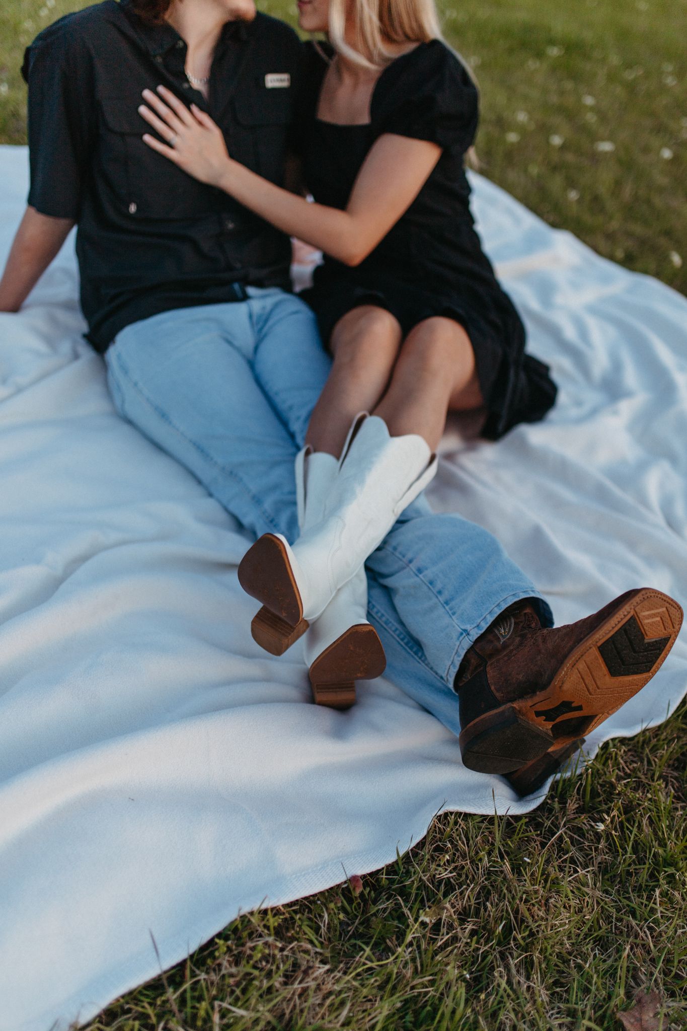 Couple sitting together on white blanket in grass wearing matching black outfits and boots.