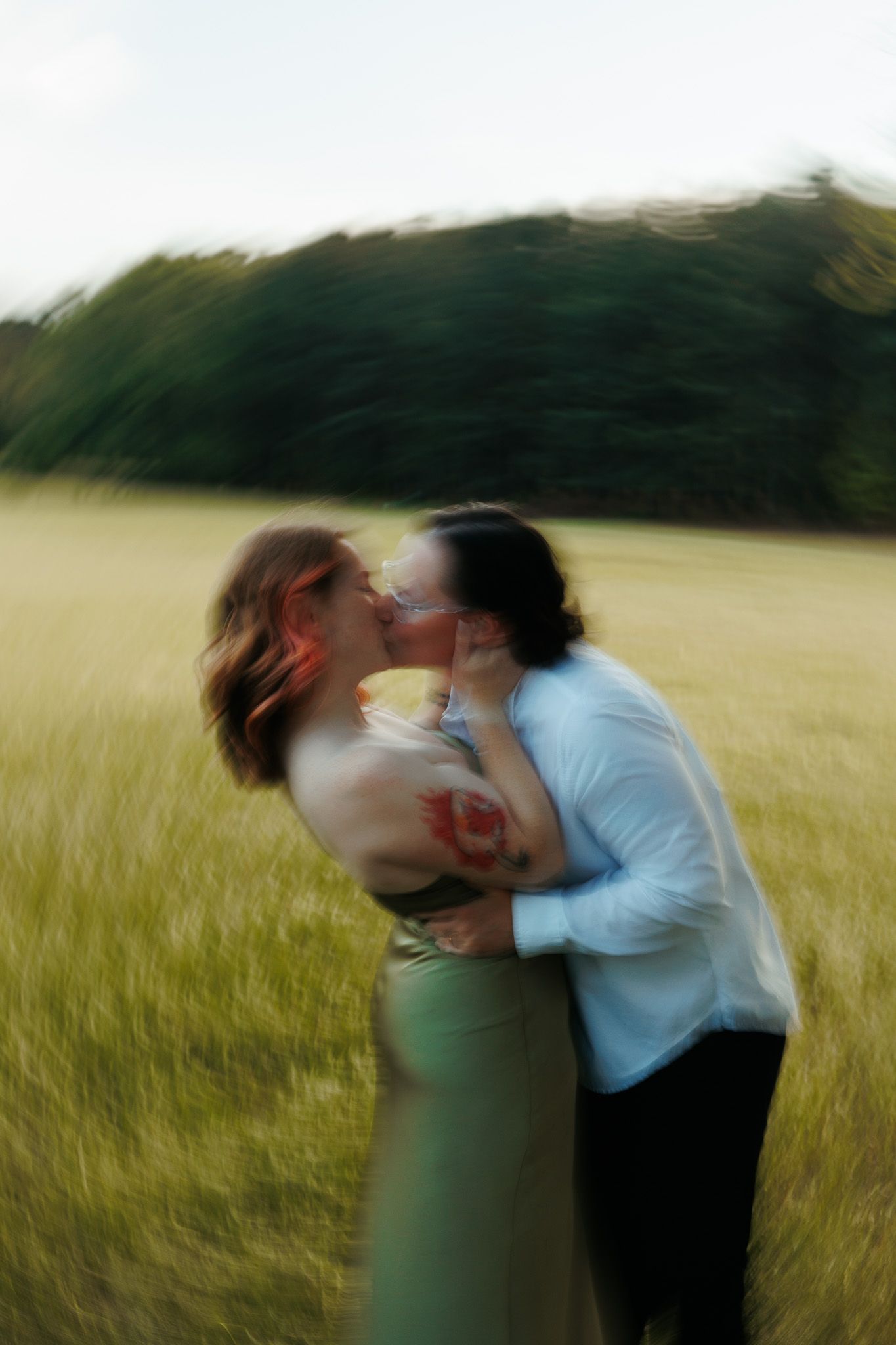 Motion-blurred photo of couple kissing passionately in green meadow with trees in background.