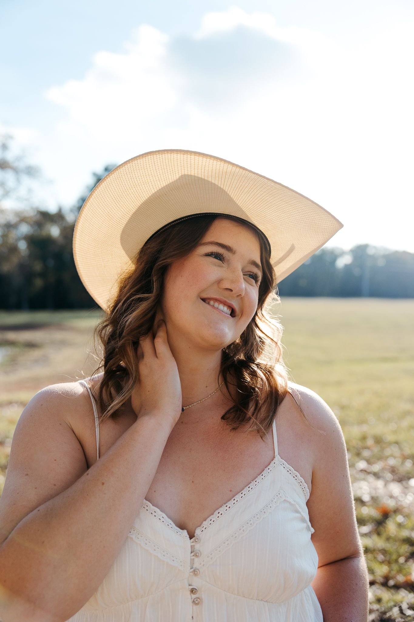 Smiling woman in white sundress and wide-brim straw hat poses in sunny field, touching her neck.