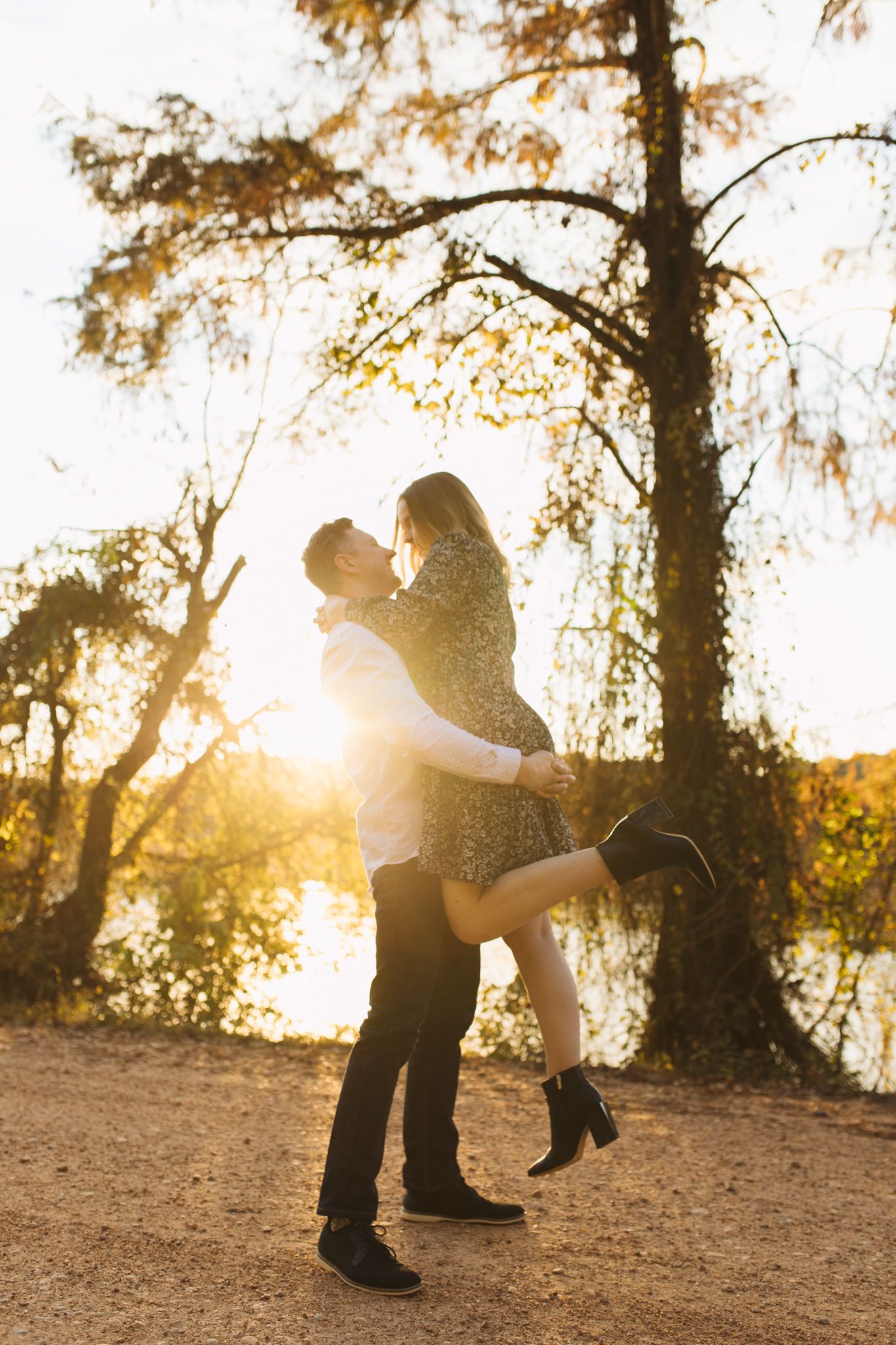 Man lifting woman in his arms near a sunlit lake surrounded by golden autumn trees.