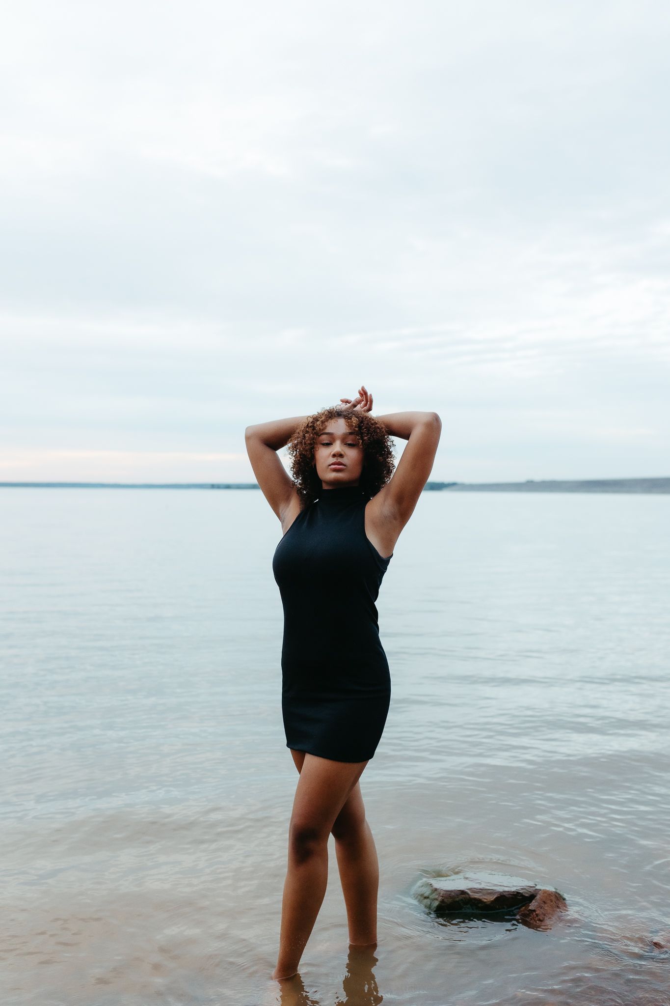 Woman in black dress posing at water's edge on overcast day, hands raised above head.