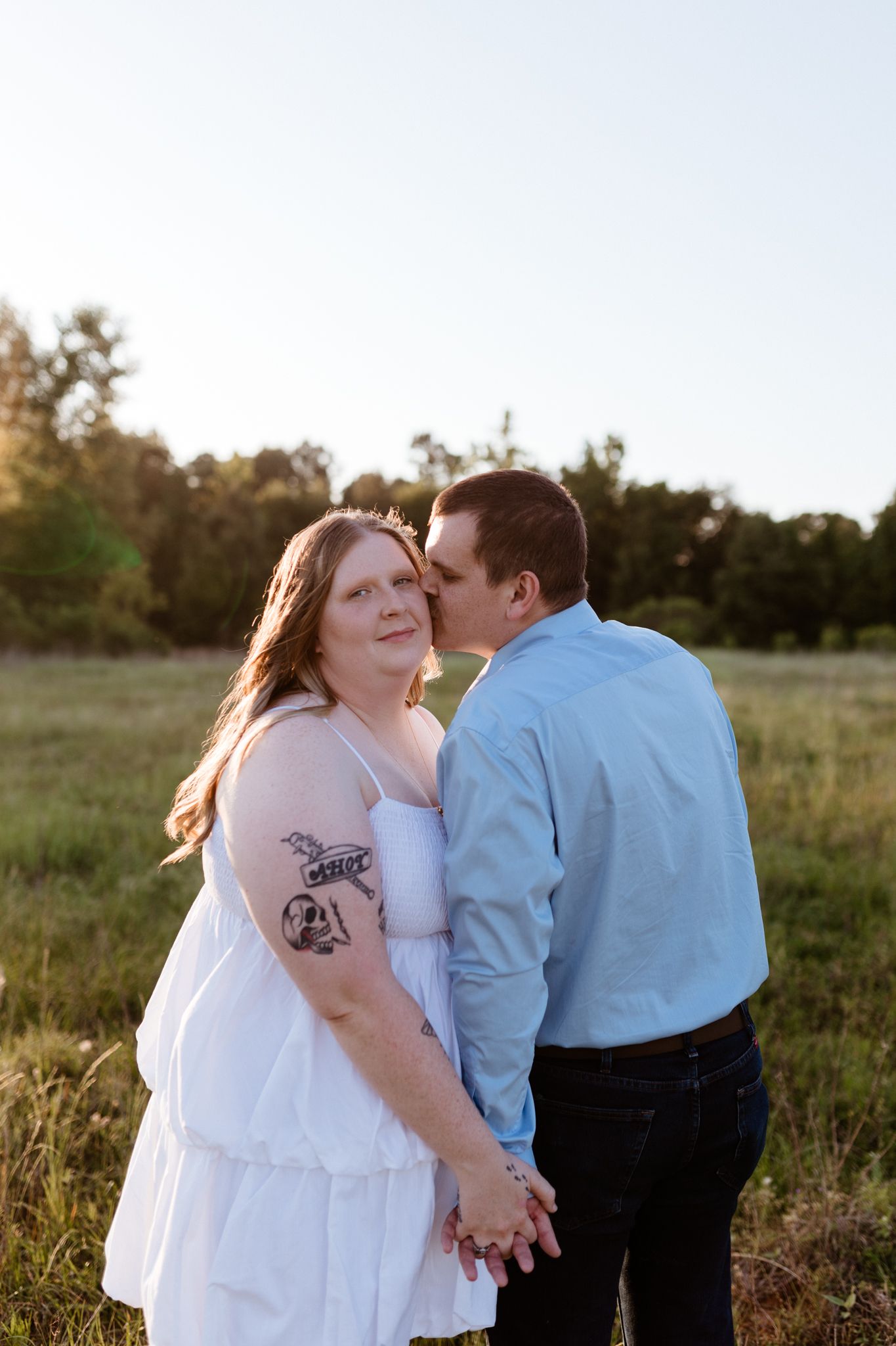 Couple holding hands in golden field, man kissing woman's cheek, she wears white dress with tattoo.