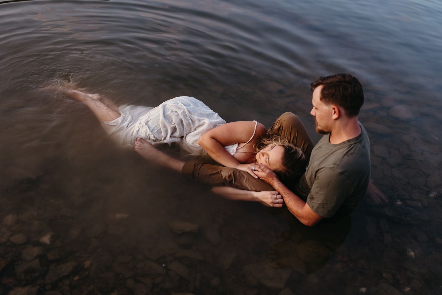 A couple shares a romantic moment in shallow water, with a woman in a white dress floating as a man holds her gently.