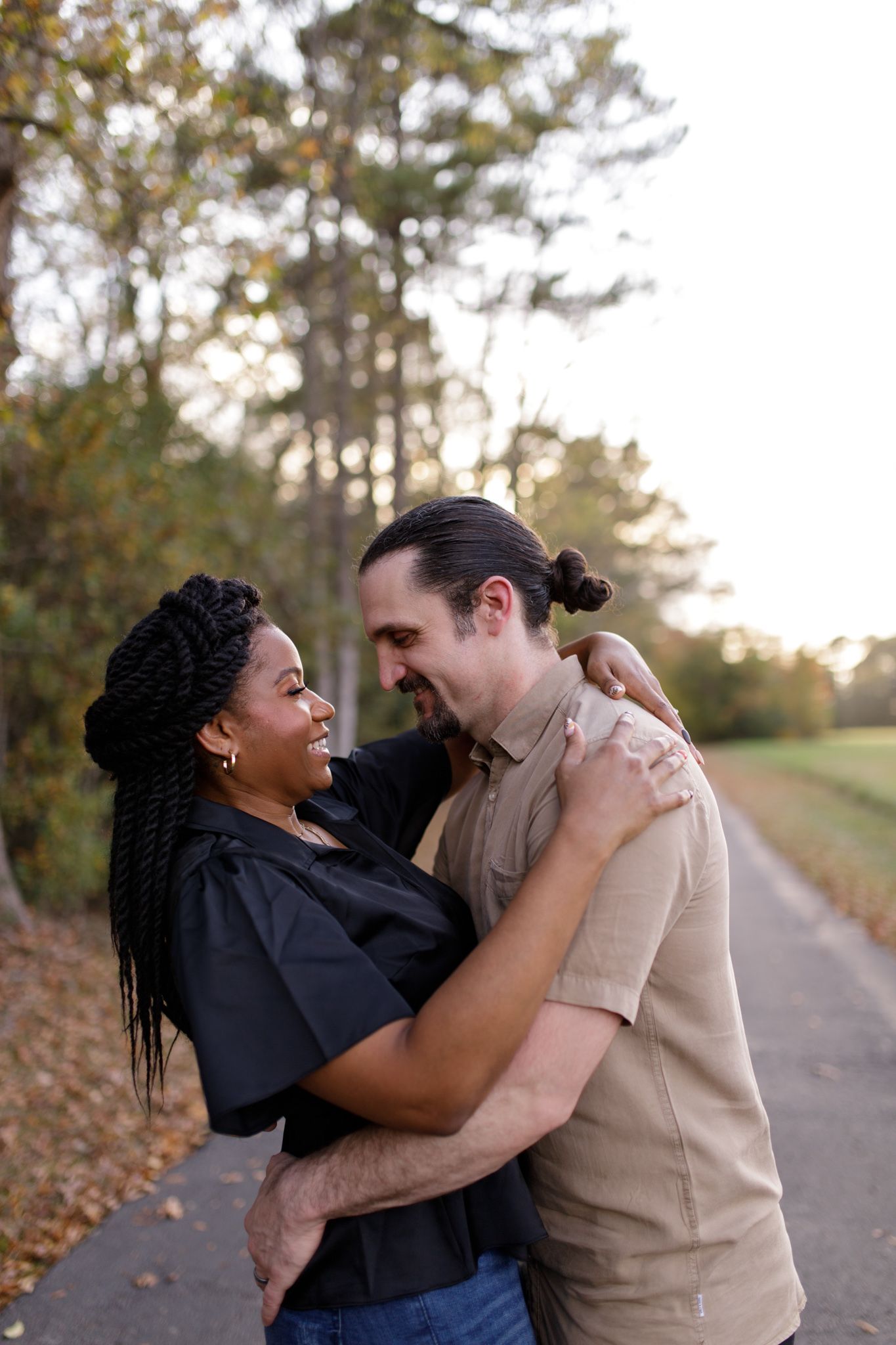 Interracial couple embracing and smiling at each other on a tree-lined path during golden hour.