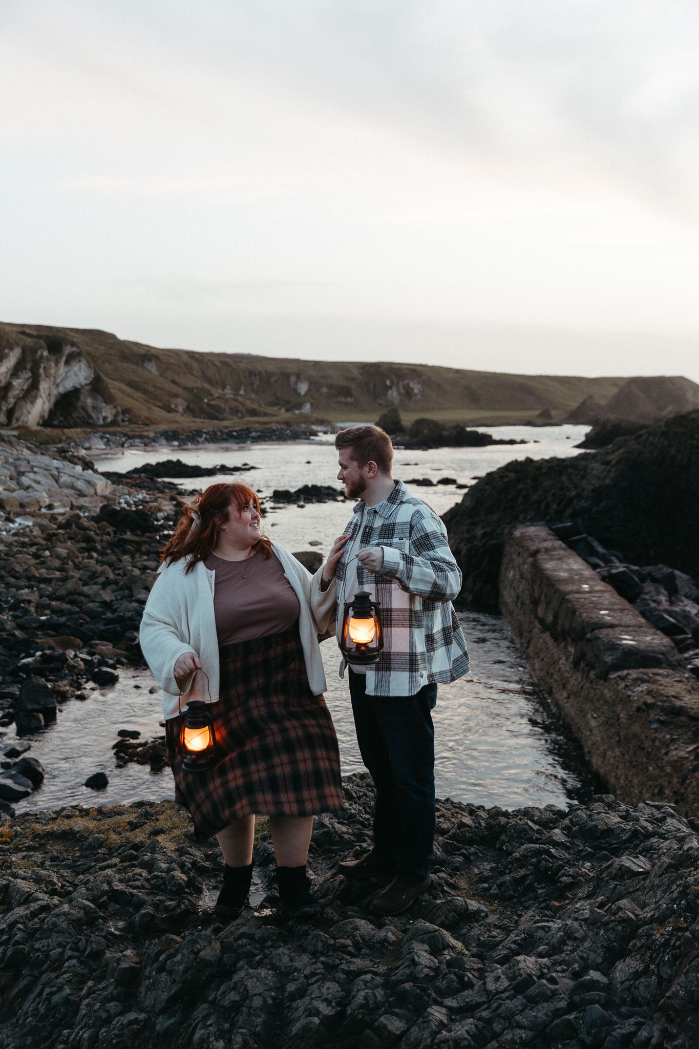 Romantic couple holding vintage lanterns on rocky coastal shoreline at dusk.