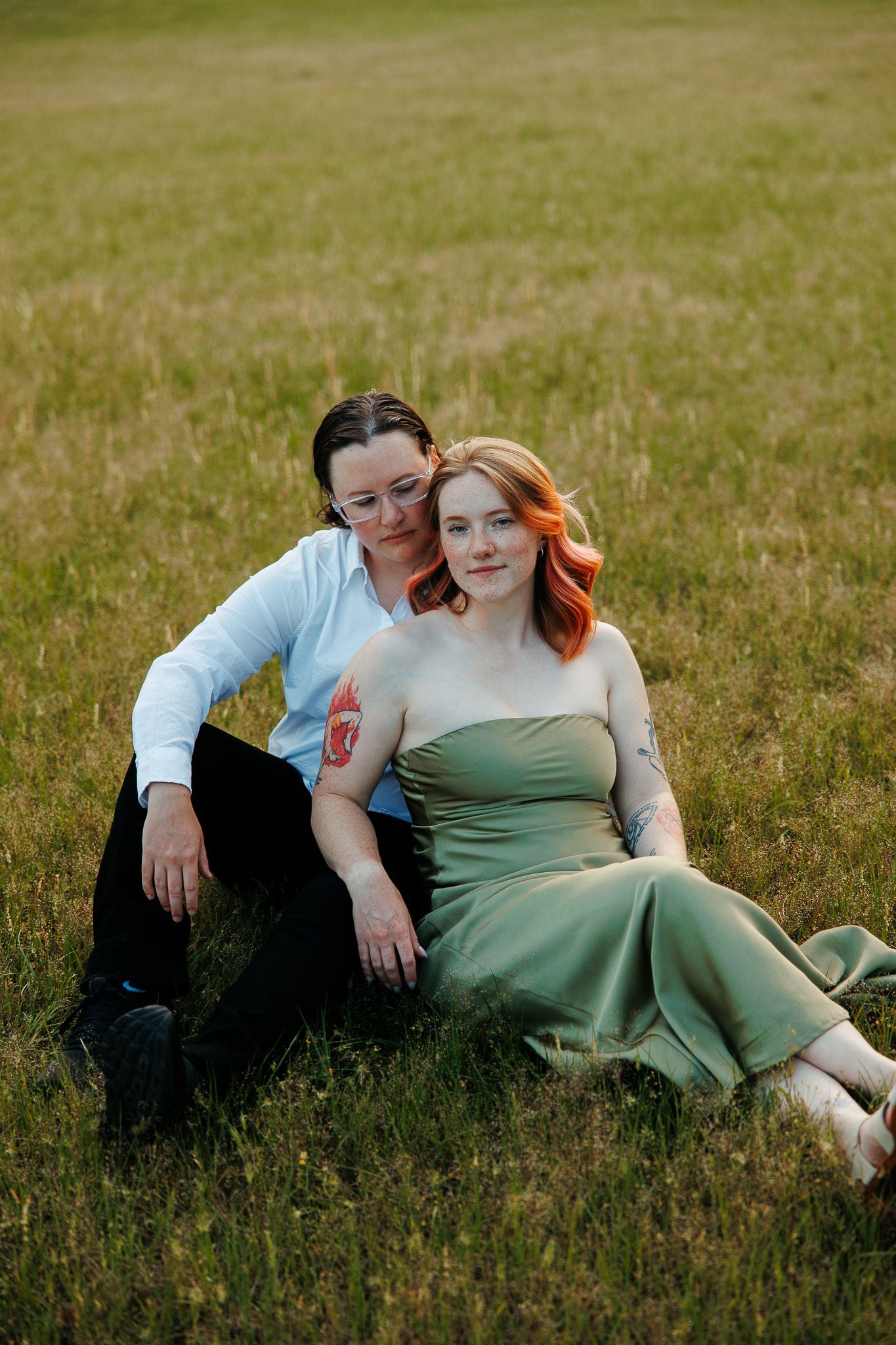 Two women sitting together in a green field, one in a white shirt and one in a sage green dress, sharing a tender moment.