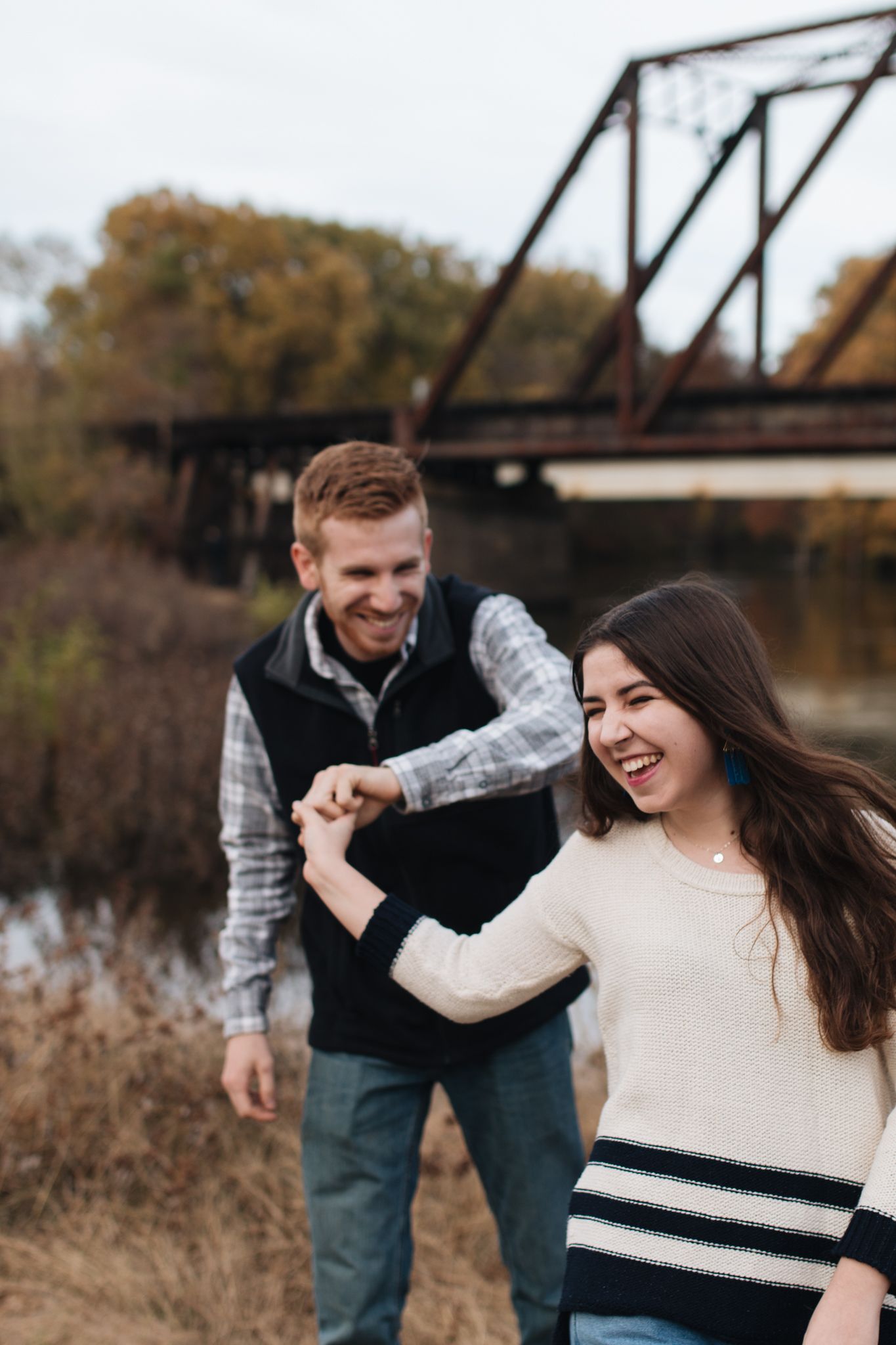 Smiling couple holding hands near a rustic iron bridge surrounded by autumn foliage.