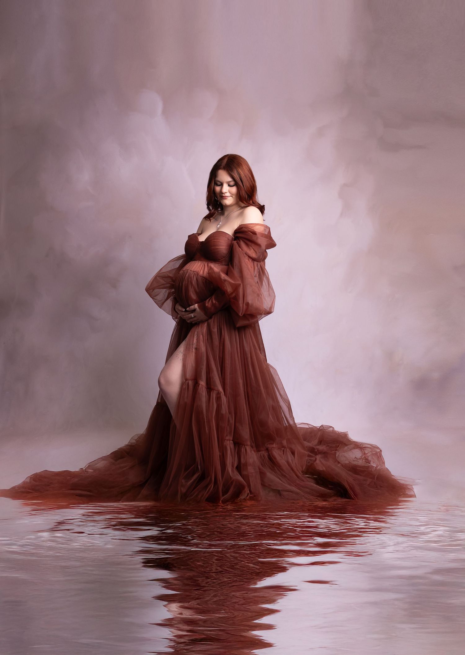Pregnant woman in flowing rust tulle gown standing in reflective water against a soft pink backdrop.