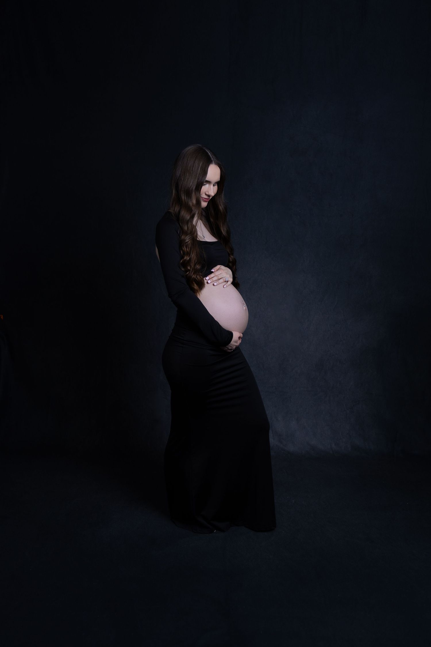 Pregnant woman in fitted black gown cradling baby bump against a dark studio background.
