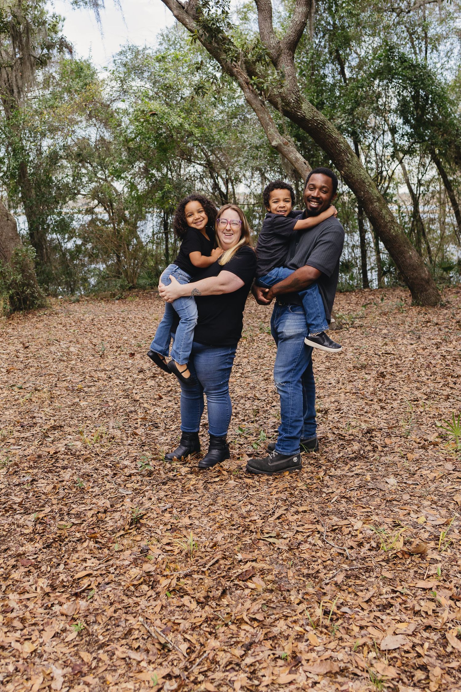 A joyful family of four posing in a forest, parents each holding one child among autumn leaves.