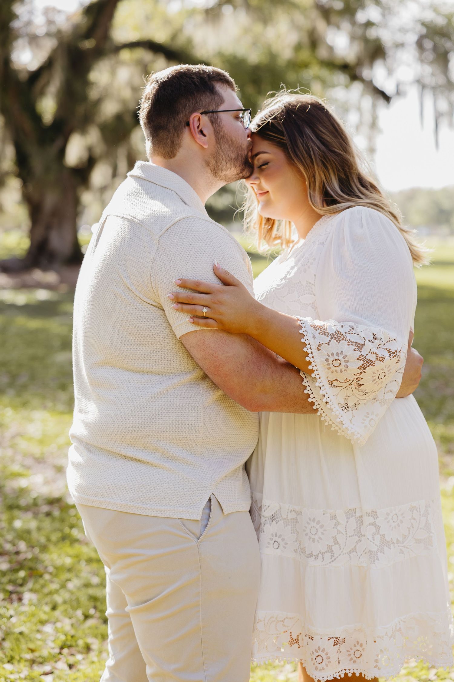 Couple sharing a tender moment outdoors, woman in white lace dress embracing man in cream outfit under mossy trees.