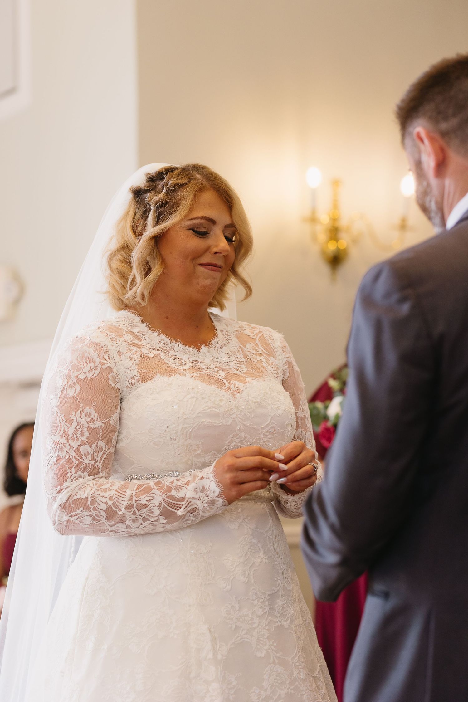 Bride in long-sleeve lace gown smiles while placing ring on groom's finger during ceremony.