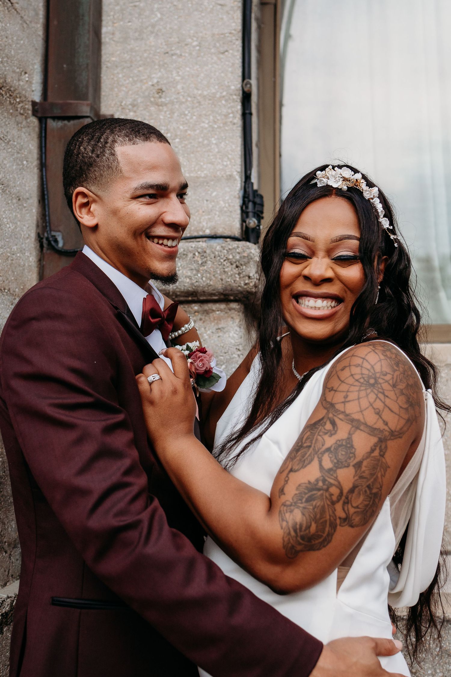 Smiling bride and groom laughing together against stone wall, bride showing dreamcatcher arm tattoo.