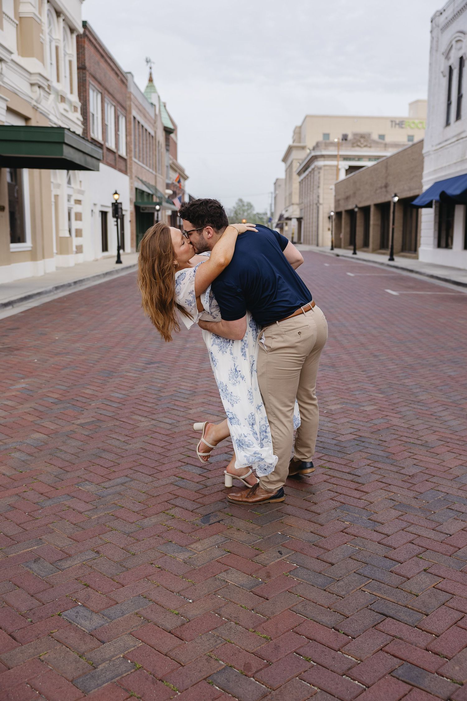 Couple sharing a romantic dip kiss on a brick pedestrian street, woman in floral maxi dress and wedge heels.