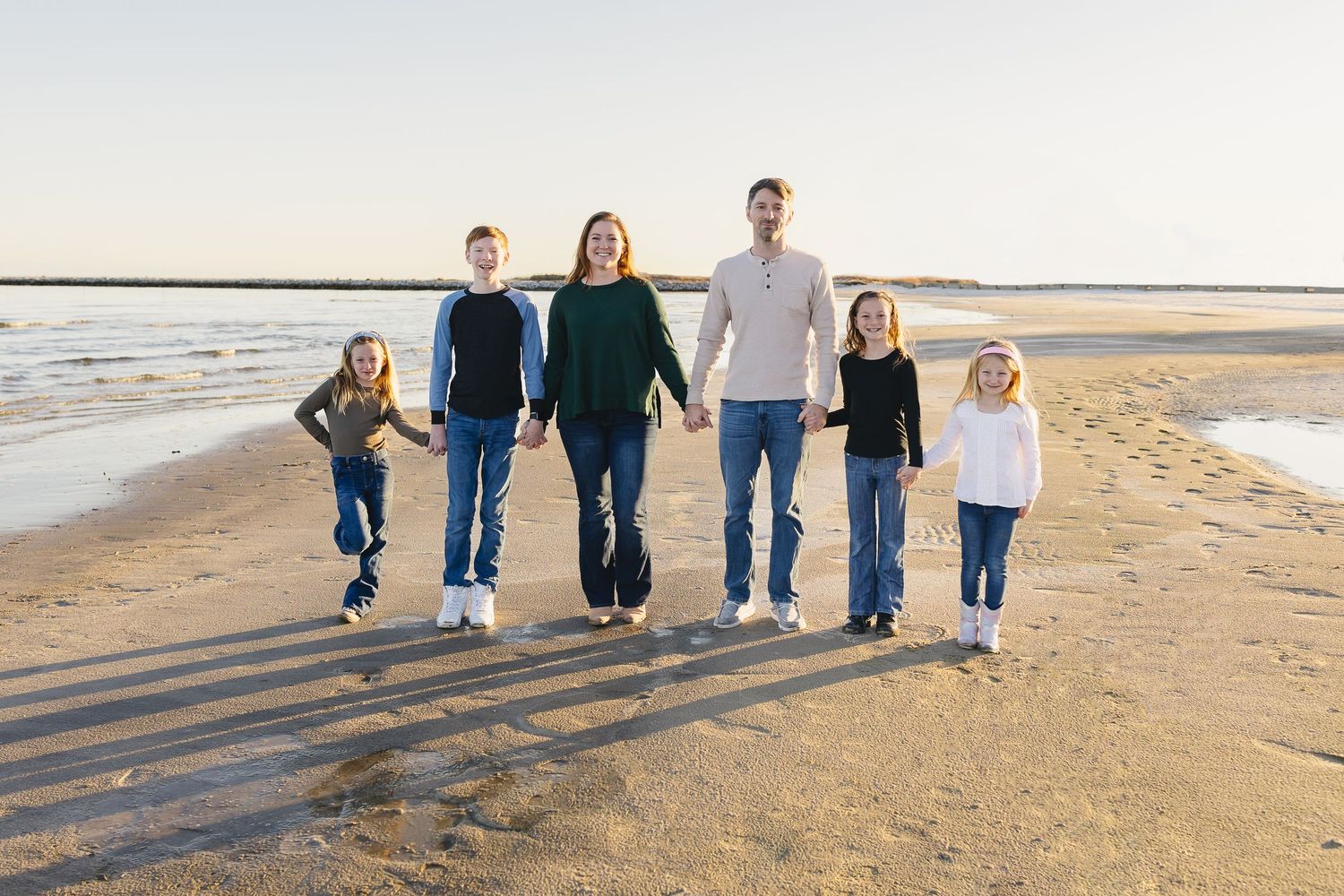 A family of six walks hand-in-hand along a wide sandy beach at golden hour.