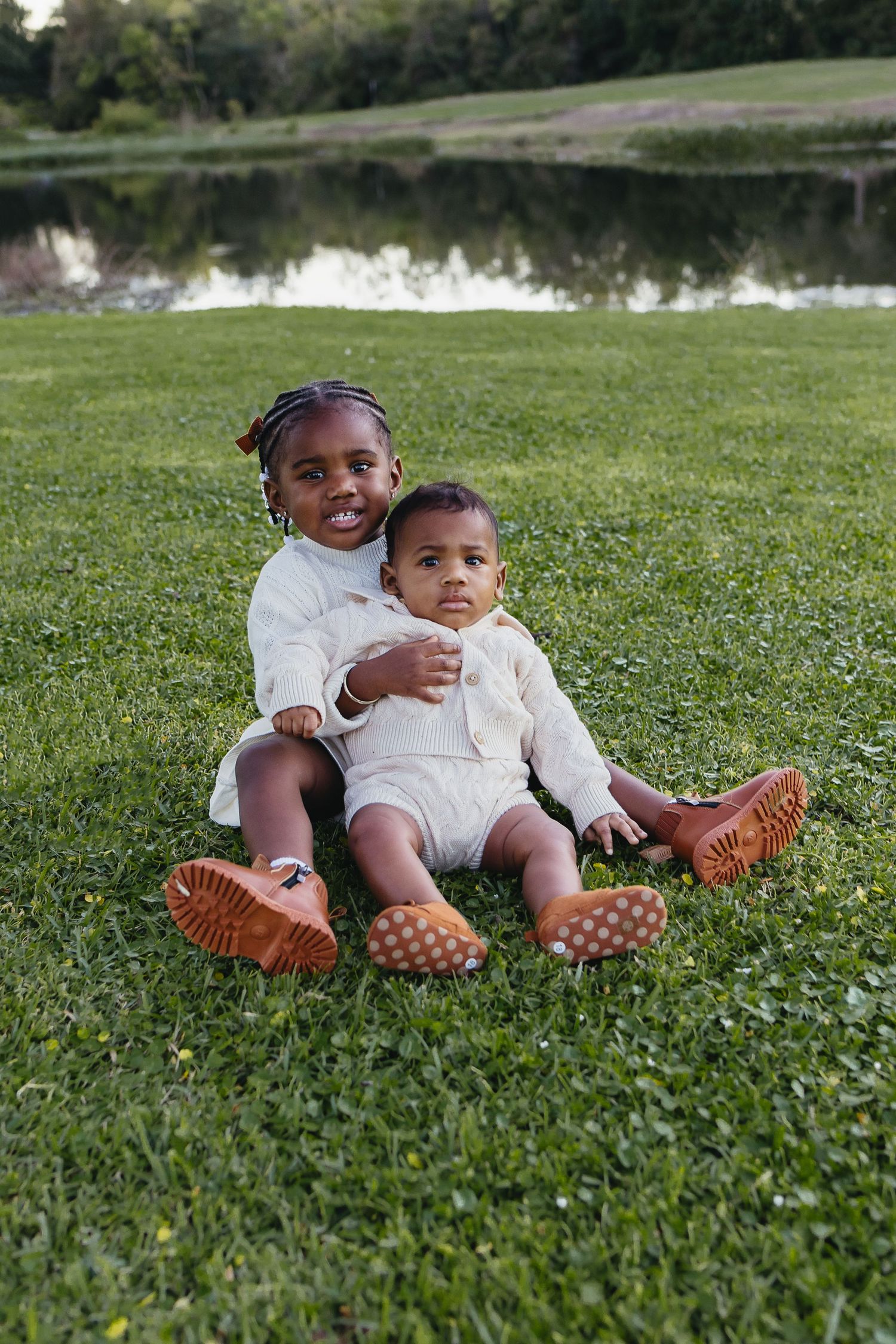 Two young Black children sitting on green grass near a pond, wearing matching cream outfits and brown shoes.