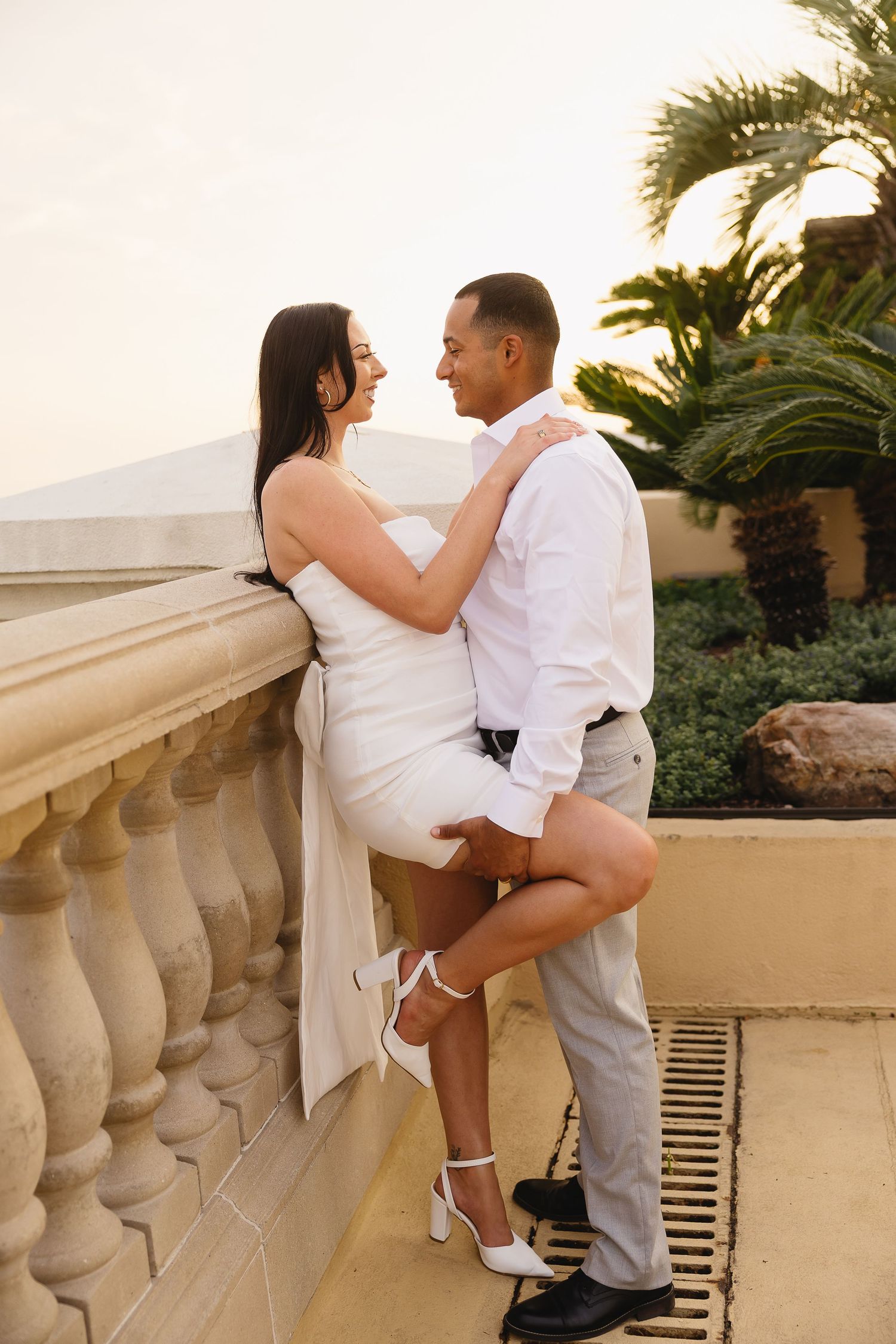 Couple laughing and embracing on balcony at golden hour, woman in white mini dress with leg raised, palm trees behind.