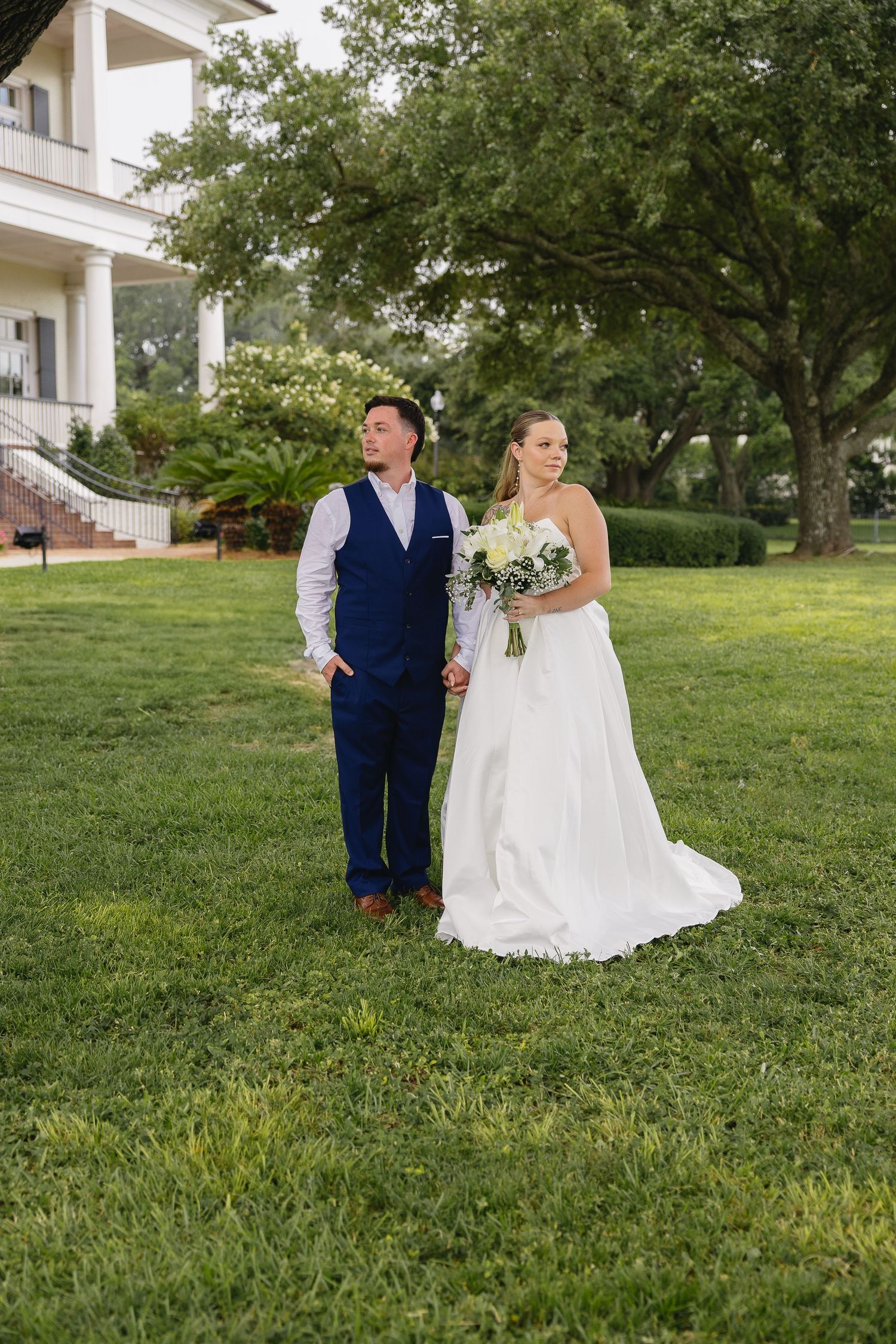 Bride holding white bouquet and groom in navy vest standing on green lawn outside white plantation venue.