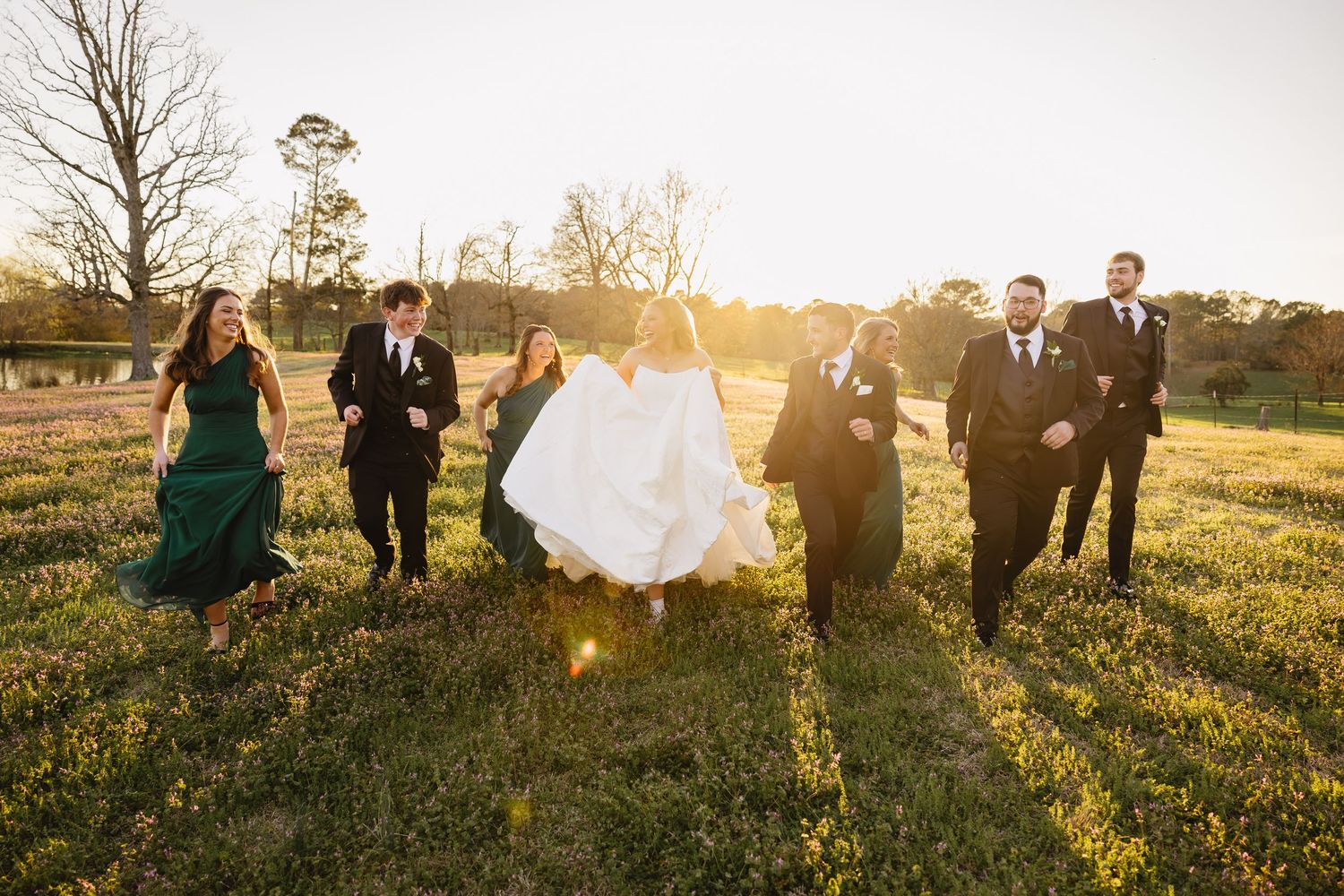 Wedding party walking together through a sunny green field at golden hour in formal attire.