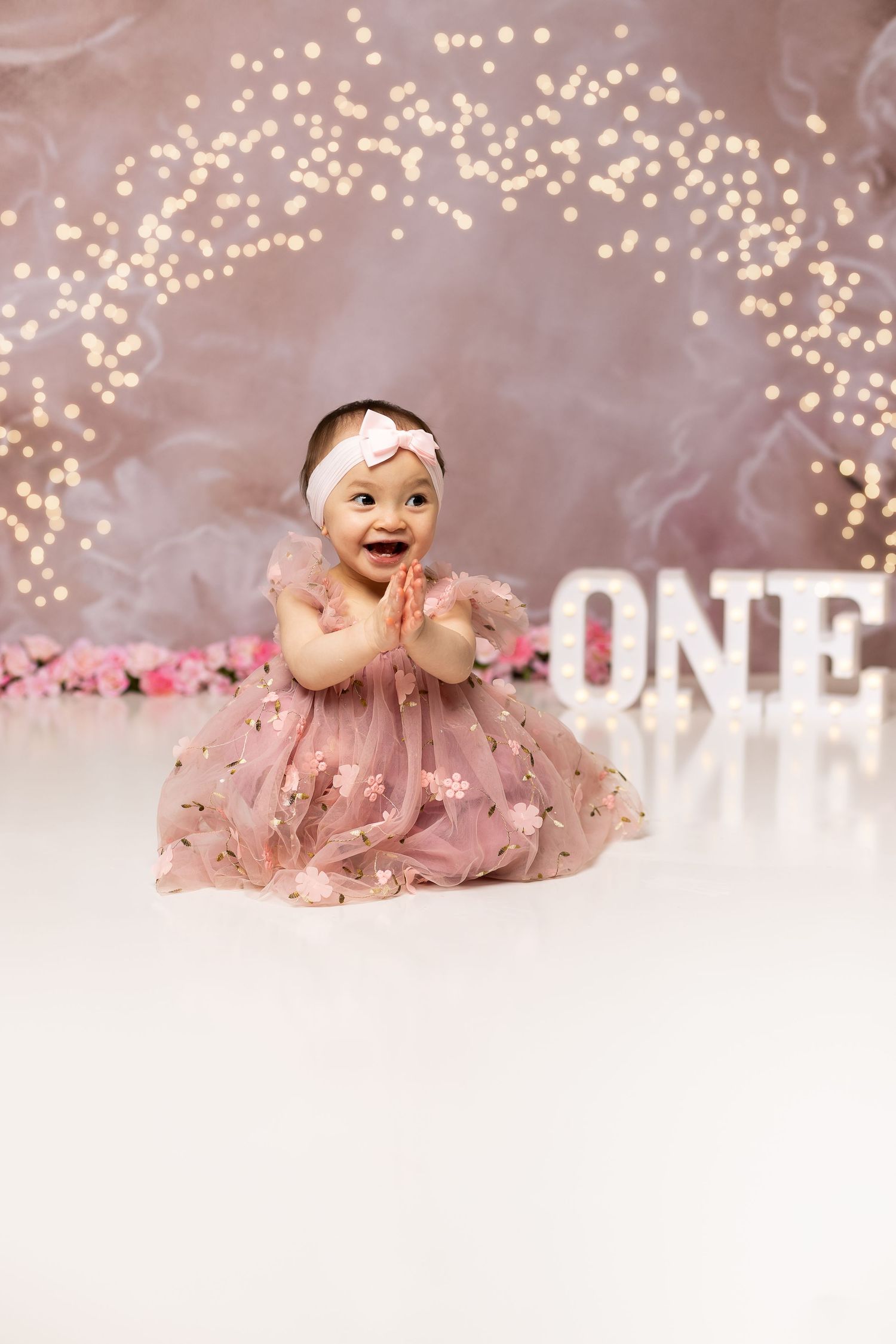 Adorable baby girl in pink dress clapping hands during first birthday cake smash photo session.