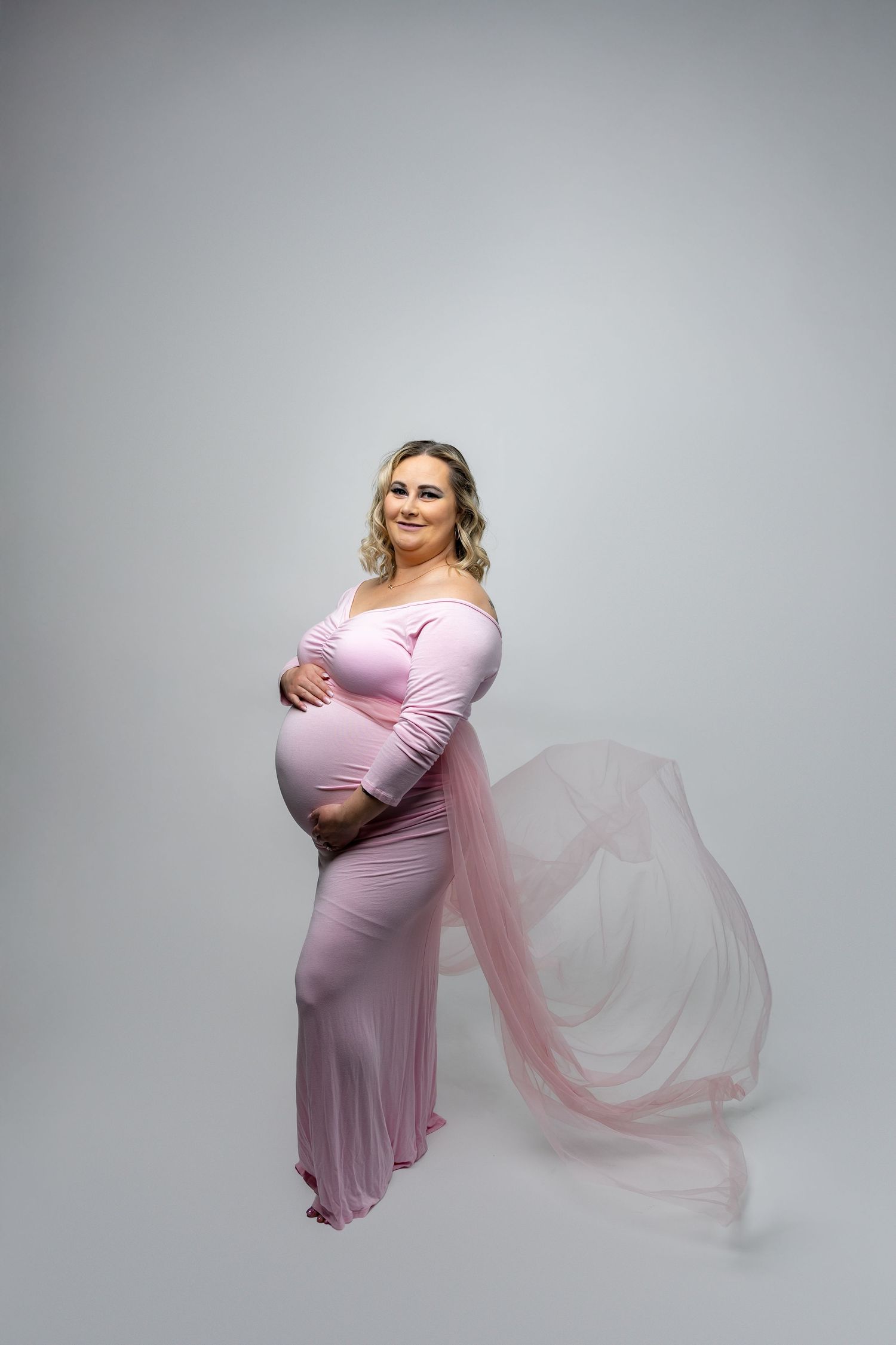 Pregnant woman in pink off-shoulder gown with flowing tulle train on white studio background.