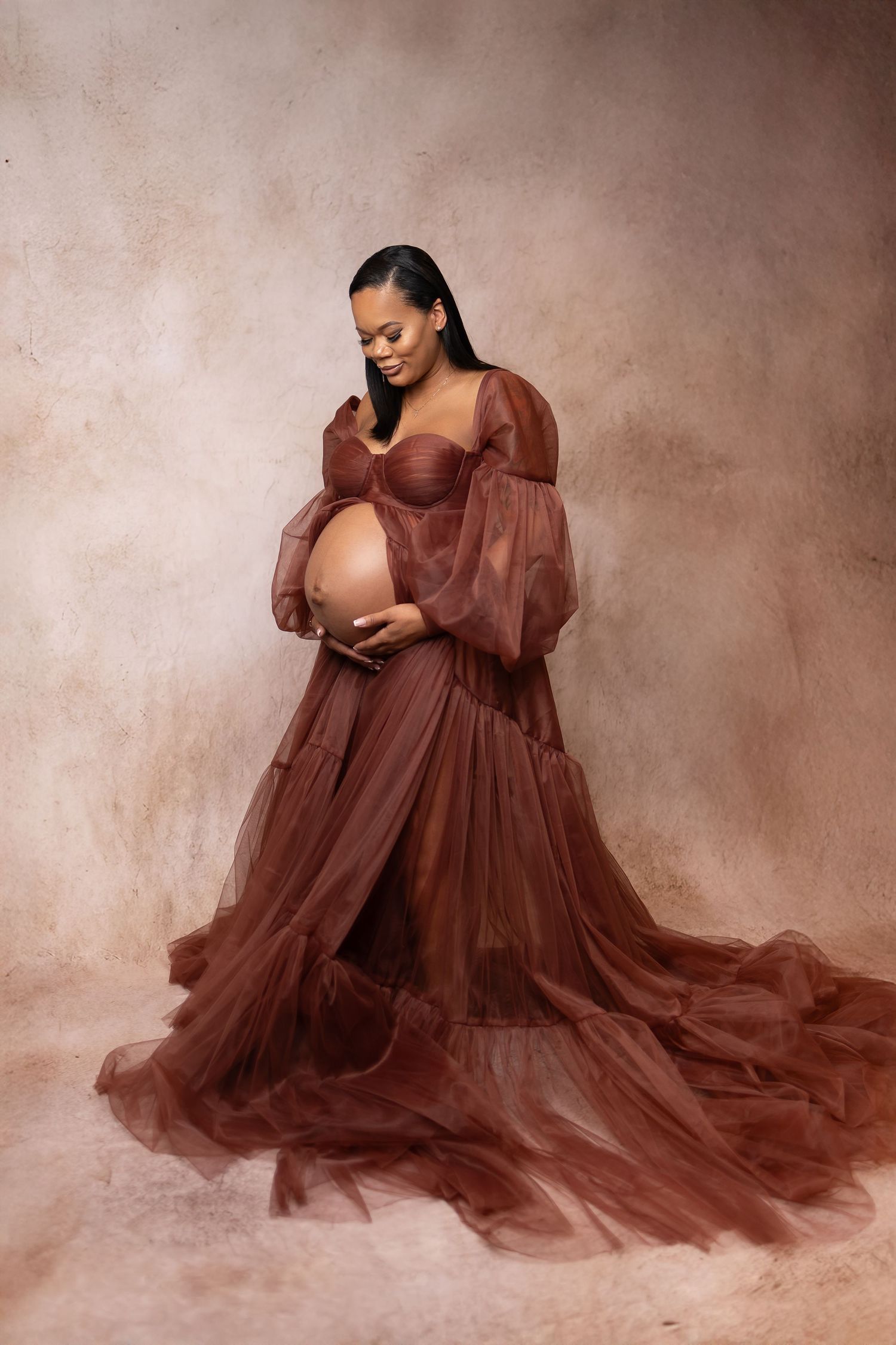 Pregnant woman in elegant brown tulle maternity gown cradling baby bump in studio portrait.